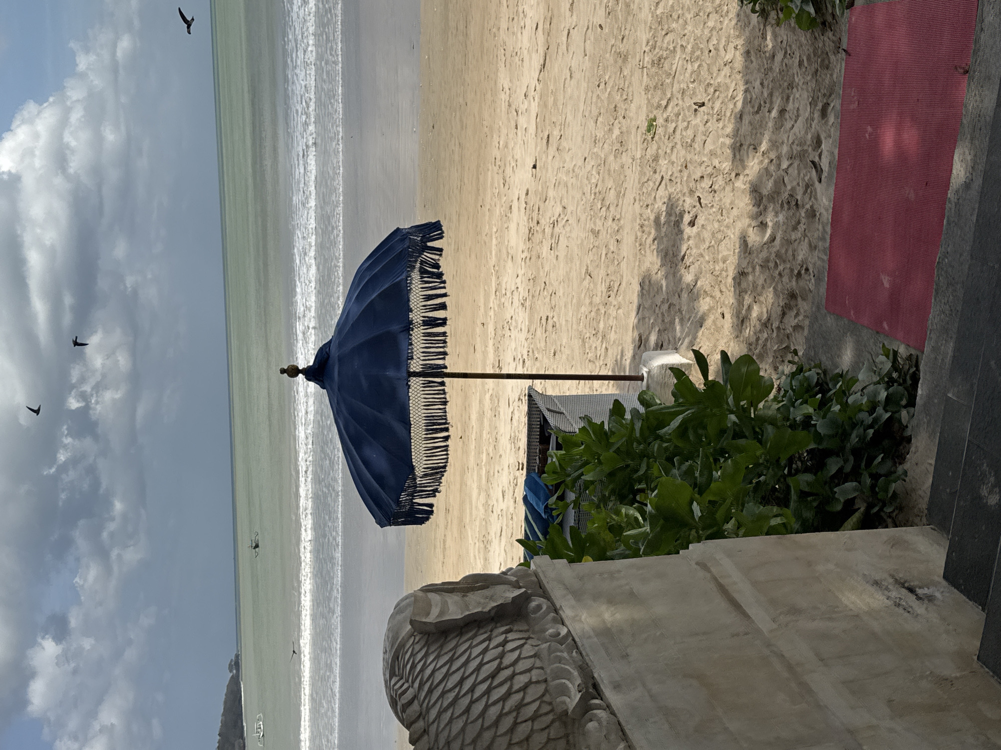 This is a medium shot image of a beach scene in Kuta Selatan, Indonesia. In the foreground, on the left, is a portion of a concrete structure with a textured surface, possibly a wall or a statue base, with dark grey steps leading down to the right. Adjacent to the concrete structure and steps, lush green foliage is visible. A red mat is placed on the sand to the right of the steps.
The middle ground of the image is dominated by a sandy beach. A dark blue umbrella with fringe detail stands open, casting a shadow on the sand. Behind the umbrella, a white wicker chair with blue towels is partially visible. Further back, the ocean stretches out, with gentle waves breaking on the shore. In the distance, there are two small boats on the water. The sky above is cloudy with patches of blue, and several birds are flying. The overall impression is a serene beach setting with elements of local cultural decoration and natural elements.