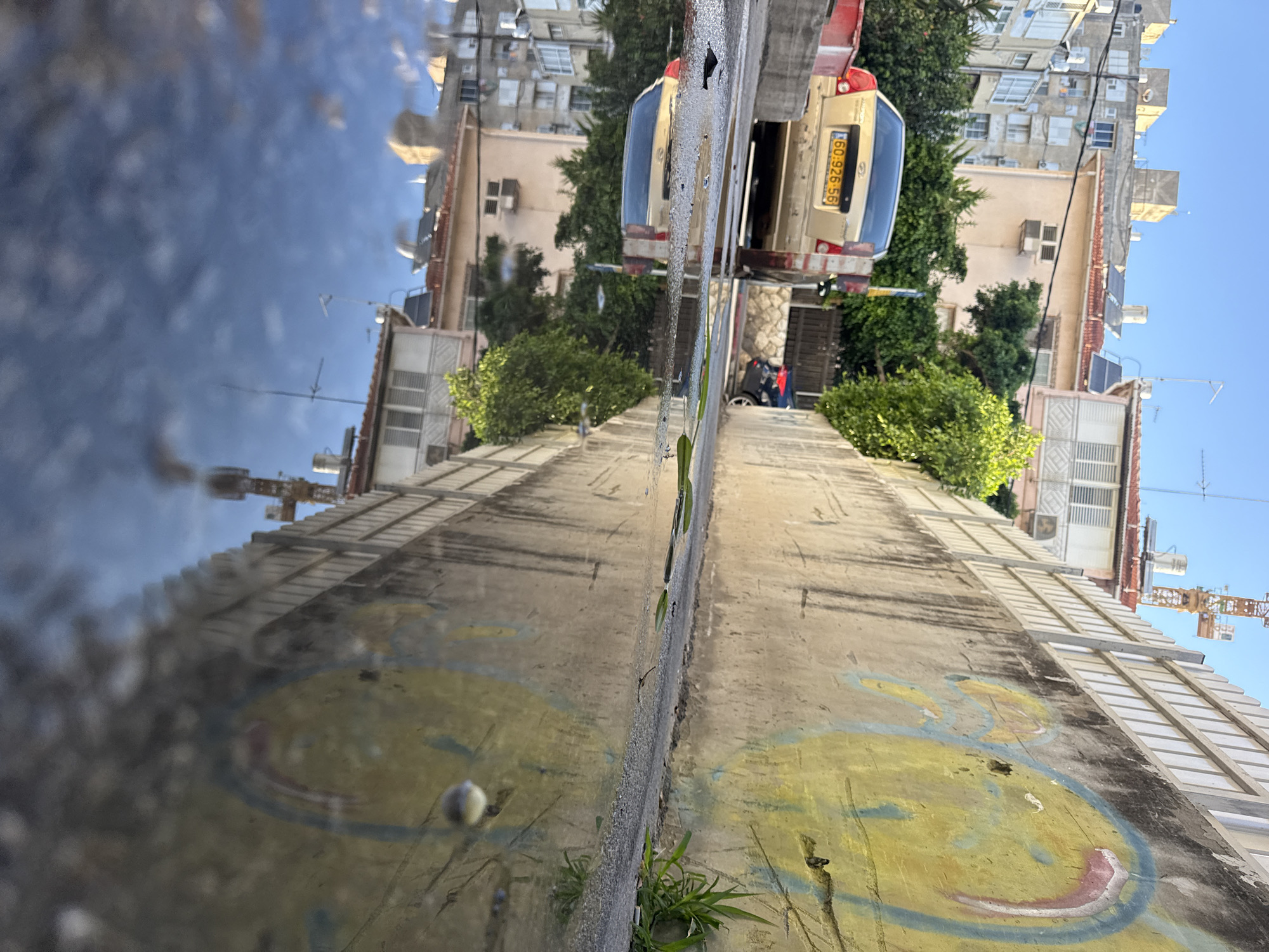 The image shows a street scene in Bat Yam, Israel, with a focus on a puddle reflecting the surrounding environment. A gold sedan with license plate 60 926 56 is parked on the left side of the street, partially obscuring the view of buildings behind it. The buildings are multi-story and appear to be residential, with some featuring balconies and air conditioning units. Lush green trees and vegetation are interspersed among the buildings.
The right side of the image is dominated by a concrete wall with a large, abstract mural painted on it in shades of yellow, blue, and pink. A portion of a white fence runs along the top of the wall. A construction crane is visible in the background, suggesting ongoing development in the area.
The puddle in the foreground creates a mirror-like reflection of the buildings, trees, and the painted wall, giving the impression of an upside-down world. The sky above is clear and blue, with bright sunlight illuminating the scene. A few blades of grass grow near the base of the wall, indicating the presence of nature in this urban setting. The overall impression is one of a typical day in a Mediterranean city, with a contrast between the man-made structures and the natural elements.