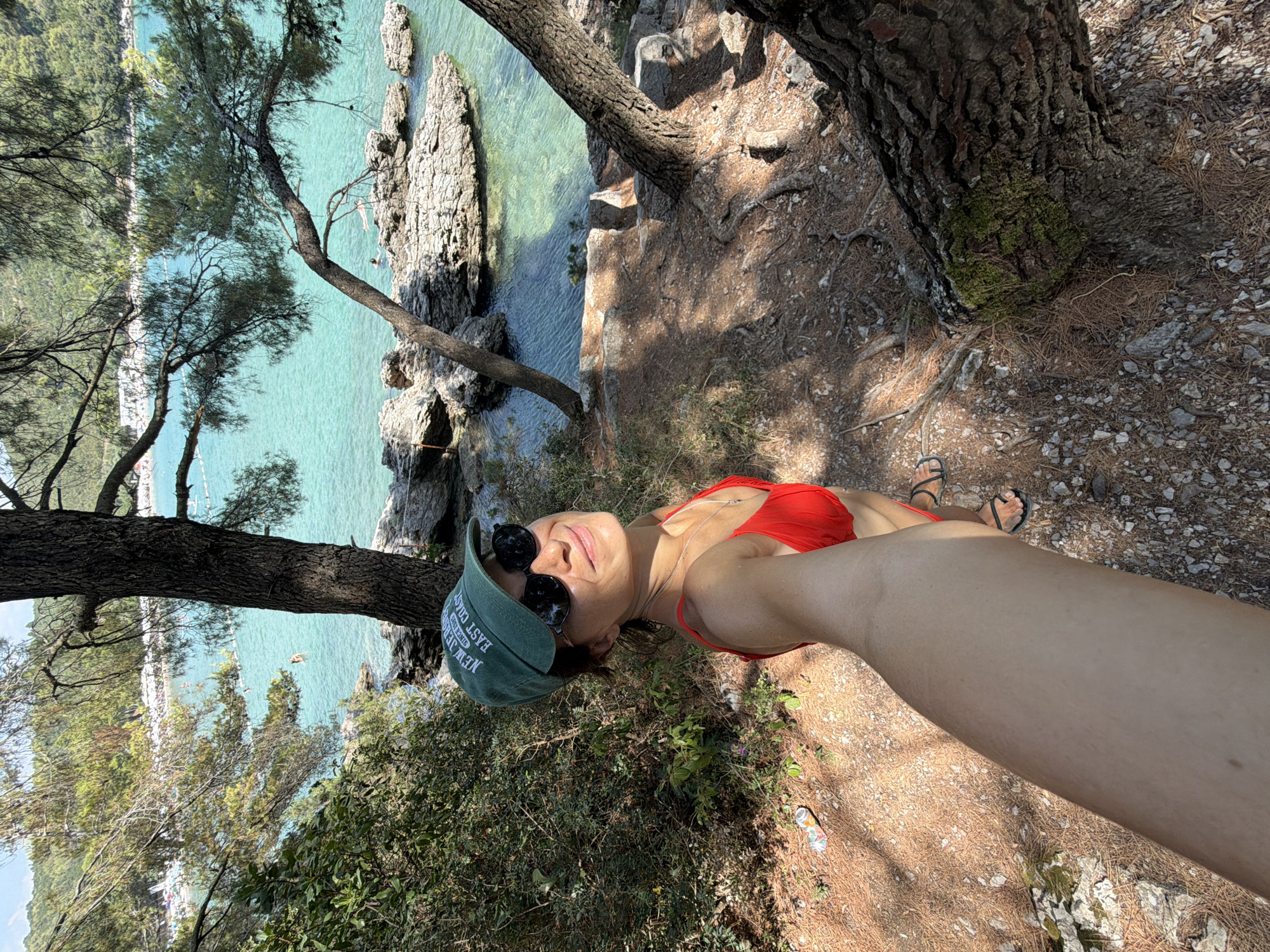 A woman is taking a selfie in Bigova, Montenegro. She is wearing a red bikini, a green baseball cap, and sunglasses. She is standing on a rocky, tree-covered outcrop overlooking a clear turquoise bay. In the background, several people are swimming and sunbathing on a beach. The sun is shining, and the water is calm.