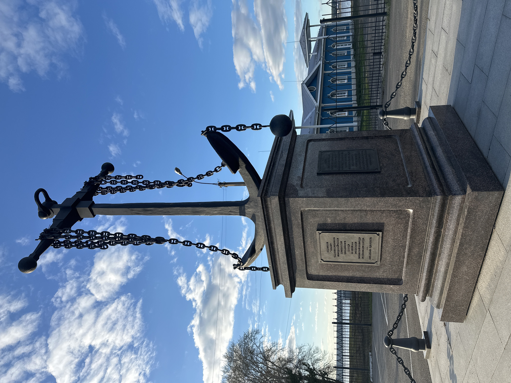 The image shows a monument in Votkinsk, Russia. It features a large anchor, draped with chains, mounted atop a granite pedestal. Two bronze plaques are affixed to the pedestal, containing inscriptions in Russian that commemorate the skills of the master craftsmen of the Votkinsk factory. The monument commemorates the 200th anniversary of the factory in September 1959. The anchor itself weighs approximately 78 tons. In the background, a blue wooden building is visible, along with a fence and a sky with scattered clouds. A bare tree is partially visible at the bottom of the image.
