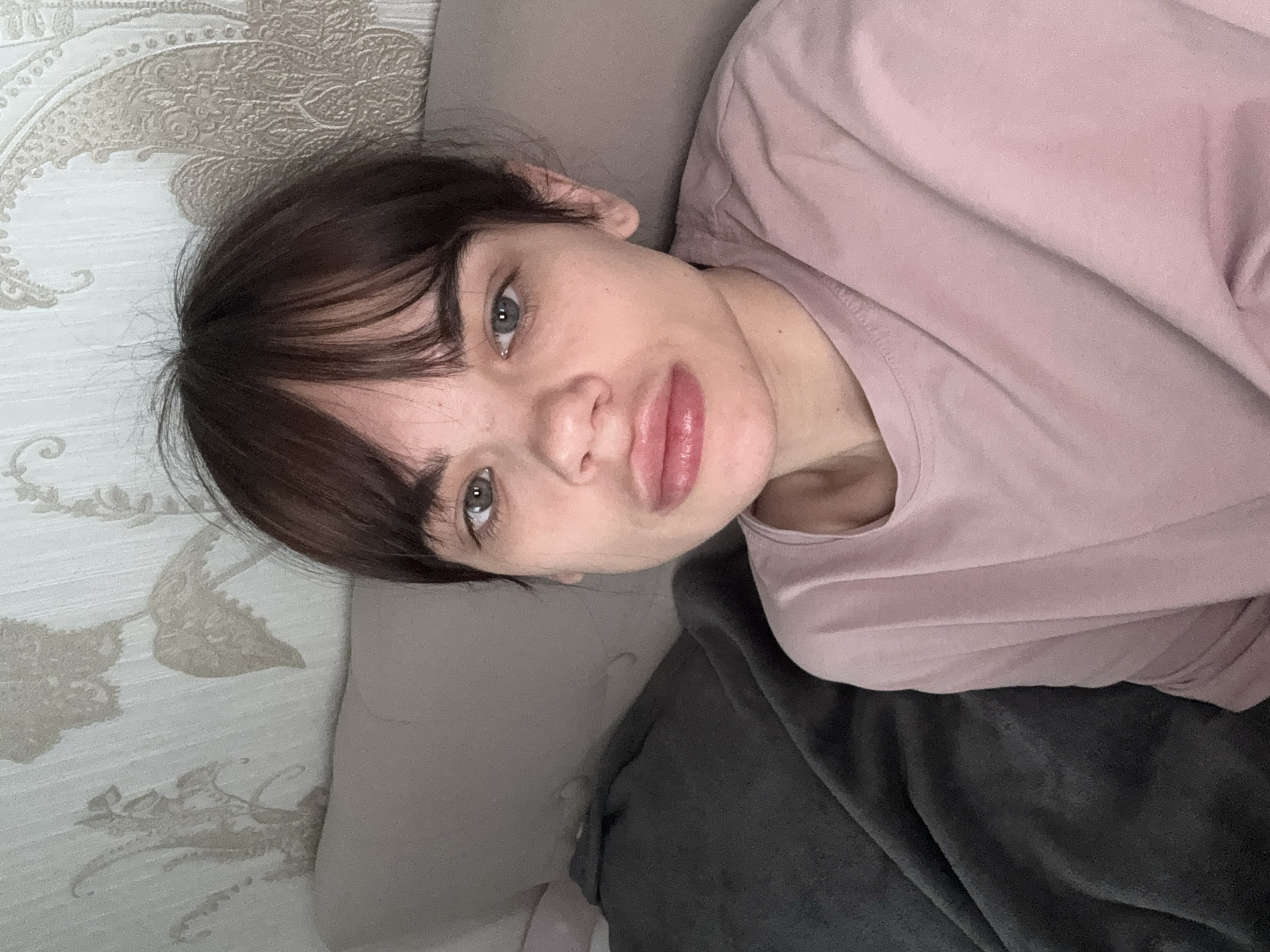 A woman lies in bed in a room with floral wallpaper. She has dark hair with bangs and is wearing a light pink t-shirt. She is resting her head on a gray pillow and partially covered with a gray blanket. The location cannot be determined to be Rostov-on-Don, Russia, based on the information in the image.