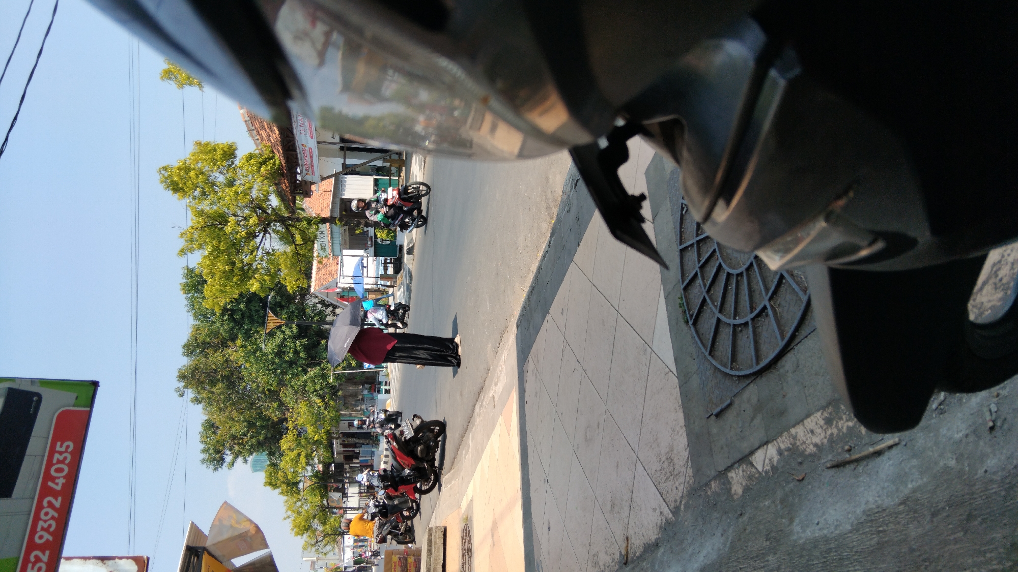 The image captures a street scene in Batang, Indonesia, on a clear, sunny day. In the foreground, the perspective is from a motorcycle, with its bodywork dominating the lower right of the frame. A circular metal grate is visible on the pavement. The street is paved, with a sidewalk to the left.
On the street, several motorcycles are in motion. To the left, a red motorcycle is parked or moving slowly, with a rider visible. Further down the street, other motorcycles are seen with riders. In the center of the image, a person stands under a black umbrella, wearing a dark maroon top and long black pants. This individual appears to be waiting or observing.
Behind the street, a row of buildings lines the road, some with tiled roofs and awnings. Trees with lush green and yellow foliage are interspersed along the street and behind the buildings, providing shade. The sky is a clear, bright blue with visible power lines stretching across the frame. A sign with red lettering, reading "52 9392 4035," is partially visible at the top left of the image. Another sign with text "TEH KING" is visible on one of the buildings. The overall impression is that of a typical bustling day on a street in a Southeast Asian city.