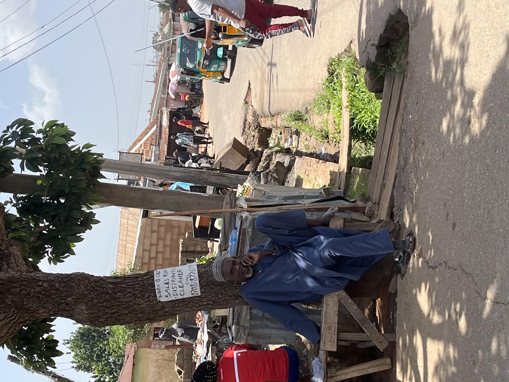 The image depicts a street scene in Jos, Nigeria, under a bright, clear sky. In the foreground, an older man with a beard, wearing a blue traditional outfit and a skullcap, is seated on a wooden bench under the shade of a large tree. A sign is attached to the tree trunk behind him, listing "WORK @ CRL SALES REP DISTRIBUTOR CLEANER" followed by a phone number.
To the left of the man, another person in a red top and dark pants is seated, their back to the camera. Various market stalls and structures with corrugated iron roofs are visible in the background, indicating a bustling commercial area. A narrow, unpaved road runs through the scene, with a damaged section featuring a hole and some vegetation growing from it.
Several people are walking or standing in the middle ground, some near market stalls, while others are on the road. Two three-wheeled auto-rickshaws, commonly known as "keke napep," are parked or driving on the road. One is green and yellow with the license plate "PTA 888," and another is primarily yellow. A man in a grey t-shirt and red pants with a stripe is standing on the right side of the road, facing away from the camera and gesturing towards the keke napep. The overall impression is one of a typical day in a Nigerian urban market environment.