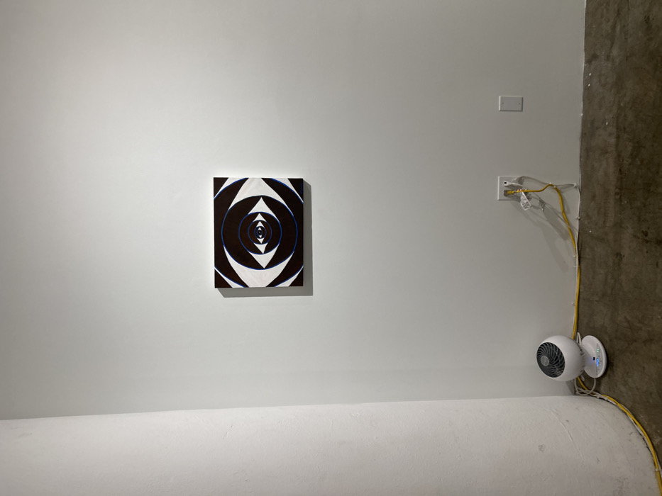This image depicts an interior space within Los Angeles, United States of America, characterized by plain white walls and a textured concrete floor. Centrally positioned on the white wall in the upper-right quadrant is a square-shaped painting. The artwork features a concentric eye motif composed of alternating black and white triangular and curvilinear shapes, with subtle blue outlines on the inner circles, creating an optical illusion of depth. On the left side of the frame, a large, round, white structural column extends vertically from the floor upwards. At the base of this column, on the concrete floor, rests a small, white, spherical Vornado-branded fan or heater, displaying illuminated blue power indicators on its front panel. A yellow cable runs along the concrete floor, secured by white clips, leading towards a duplex electrical outlet on the lower-right wall. A white power adapter is plugged into this outlet, with a segment of the yellow cable connecting to it. To the right of the active outlet, a white rectangular wall plate is mounted. The concrete floor exhibits a dark, mottled appearance with visible wear and lighter dust patches. No individuals or animate subjects are present in the scene.