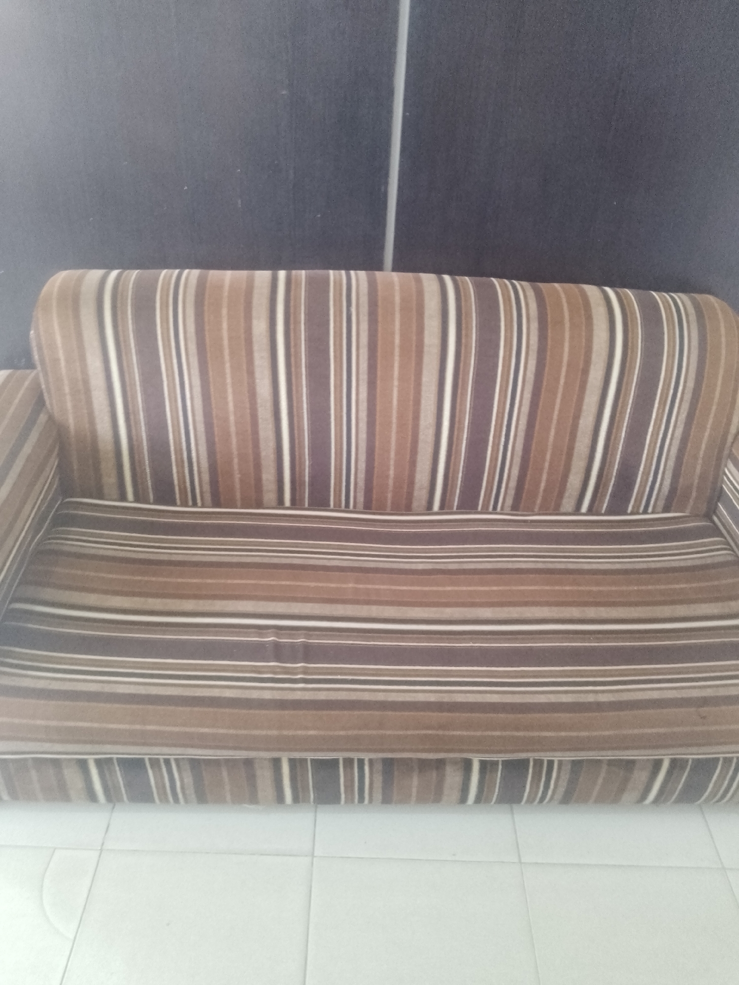 The image shows a striped sofa positioned against a dark, textured wall. The sofa has a pattern of vertical stripes in varying shades of brown, white, and dark grey on its backrest and arms. The seat cushion also features horizontal stripes in similar colors. The floor in front of the sofa is tiled in a light color, possibly white, with visible grout lines forming a grid. The overall scene appears to be indoors, likely in a residential setting. There is no indication of people or any specific actions taking place. The location in Gura Top, Nigeria is not visually discernible from the image itself.