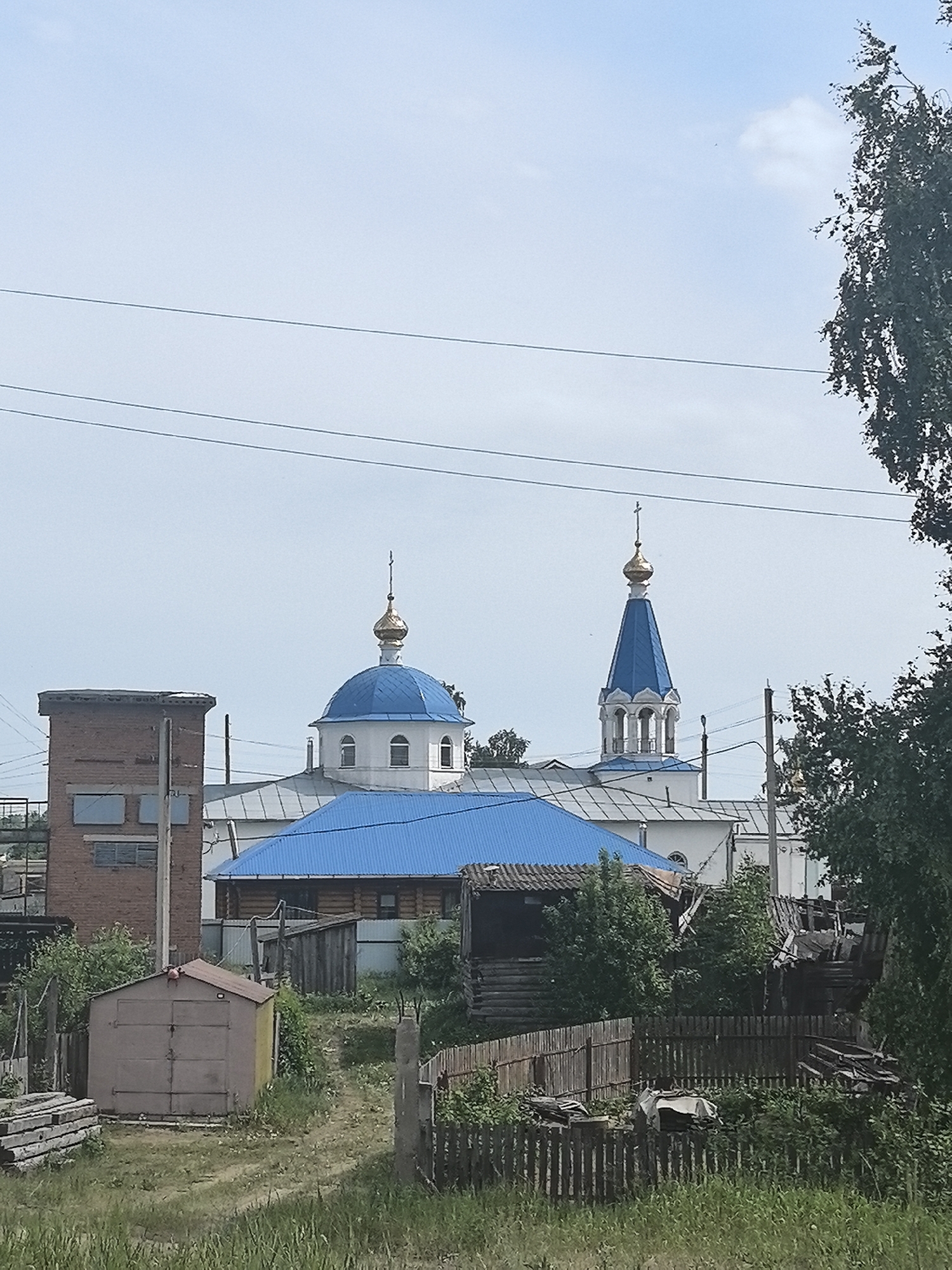 The image shows a view of the Church of the Intercession of the Holy Virgin in Noviy, Russia, behind a residential area. The church features a white exterior with blue domes topped with gold crosses. In the foreground, there's a mix of structures, including a brick building, wooden houses with blue roofs, a small shed, and a wooden fence. The ground is covered in grass and scattered debris. Overhead, power lines cross the sky, and a tree occupies the right edge of the frame.