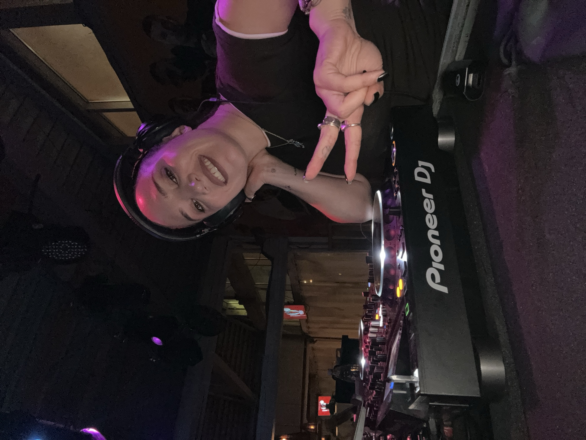 A DJ stands in front of a Pioneer DJ mixing console, illuminated by its multicolored lights. She wears headphones and makes a peace sign with her right hand, smiling toward the camera. A black t-shirt is visible, along with rings on her fingers. The background appears to be a dimly lit interior of a building, potentially a club or event space. The location is possibly in Moscow, Russia, based on structural elements and atmosphere resembling local venues.