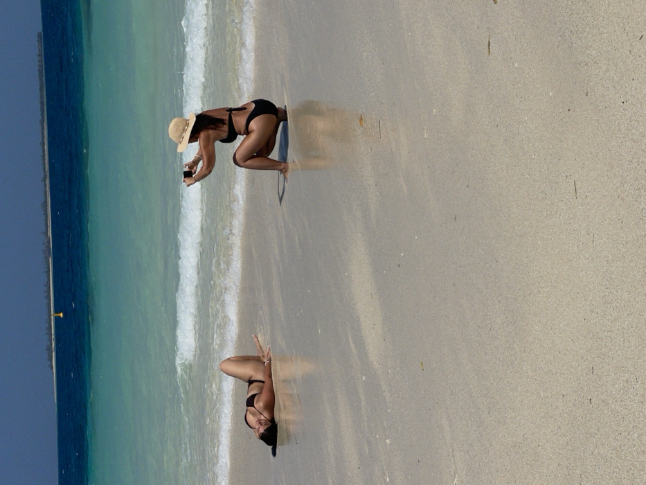 Two women on a sandy beach bordering turquoise water. The woman at the top wears a bikini and straw hat, crouching to take a photo with her phone. Her reflection is visible in the sand. The second woman, closer to the water's edge, also wears a bikini and is possibly striking a pose. The sky above the ocean is a clear blue.