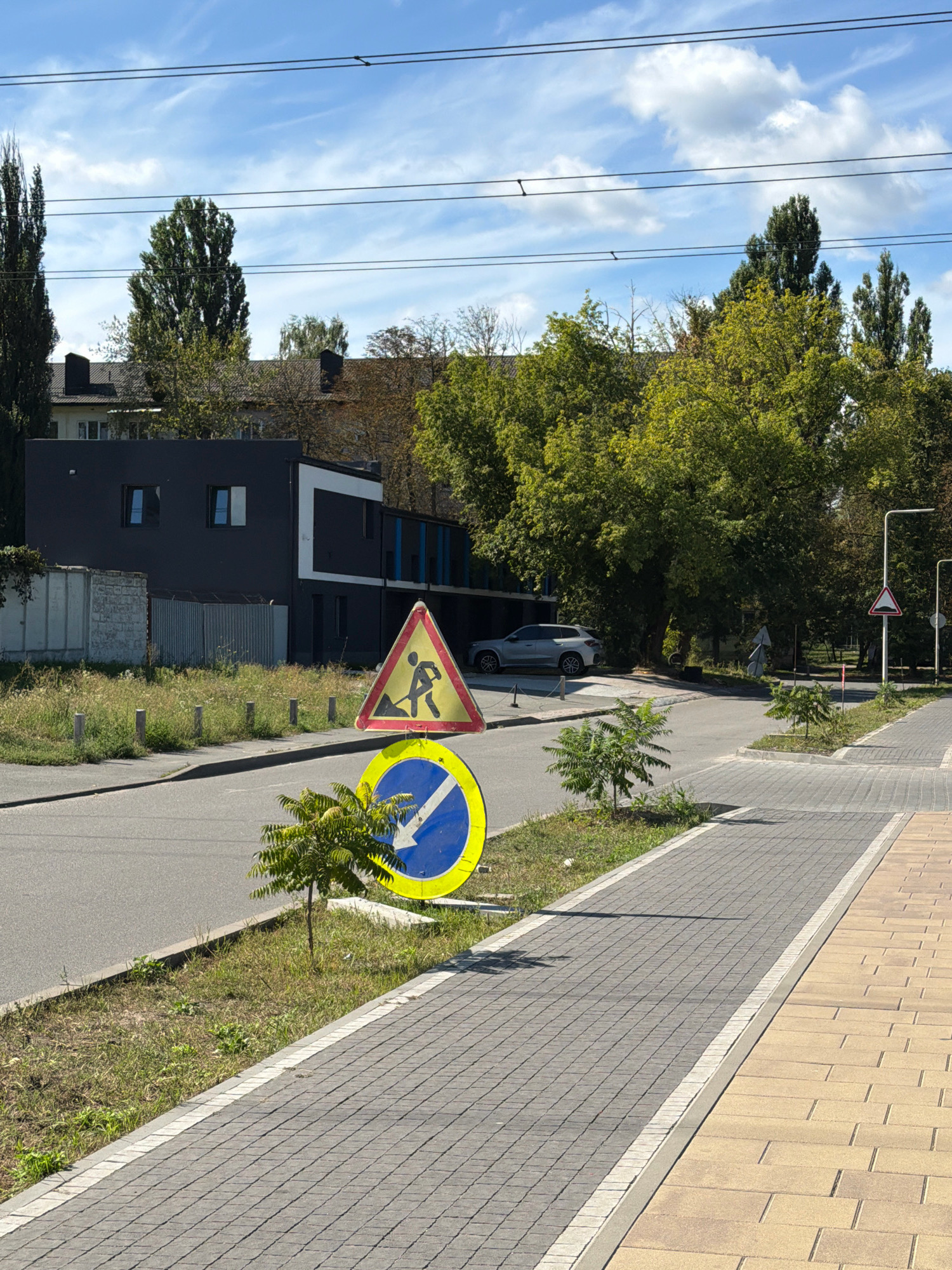 The image depicts a street scene on a clear day in Kyiv, Ukraine. In the foreground, a grey paved sidewalk runs alongside a road. A small tree is planted in a grassy strip between the sidewalk and the road. Two traffic signs are visible: a yellow triangular sign with a black silhouette of a person digging, indicating road work, and a blue circular sign with a white arrow pointing downwards and to the left, suggesting a mandatory direction of travel. Further down the road, a silver SUV is parked. Behind the road, a modern, dark-colored building with white and blue accents stands amidst lush green trees. Power lines stretch across the sky, and a few white clouds are present. The overall environment suggests an urban or suburban setting with ongoing construction or maintenance.