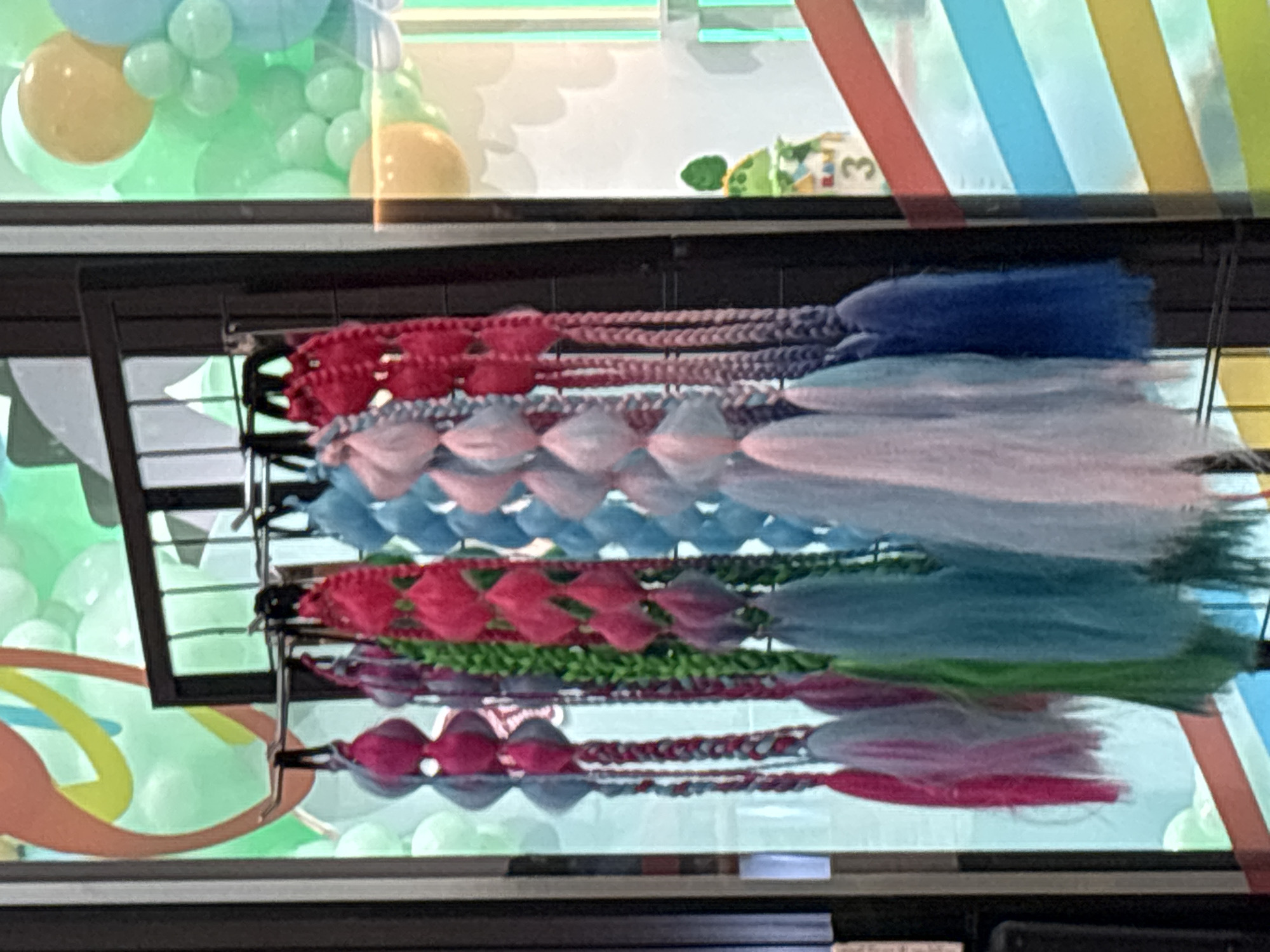 A close-up, eye-level shot captures a rack of colorful, braided hair extensions displayed behind a glass window, likely in a shop or market in Dubai, United Arab Emirates. The extensions, varying in shades of pink, blue, green, and purple, are hung from a metal rack. Each extension features a series of bulbous, colored beads separated by braided strands, culminating in a tassel-like fringe. In the background, out of focus, are balloons and a colorful, diagonal stripe pattern, suggesting a festive or party supply store. The overall scene is bright and vibrant.