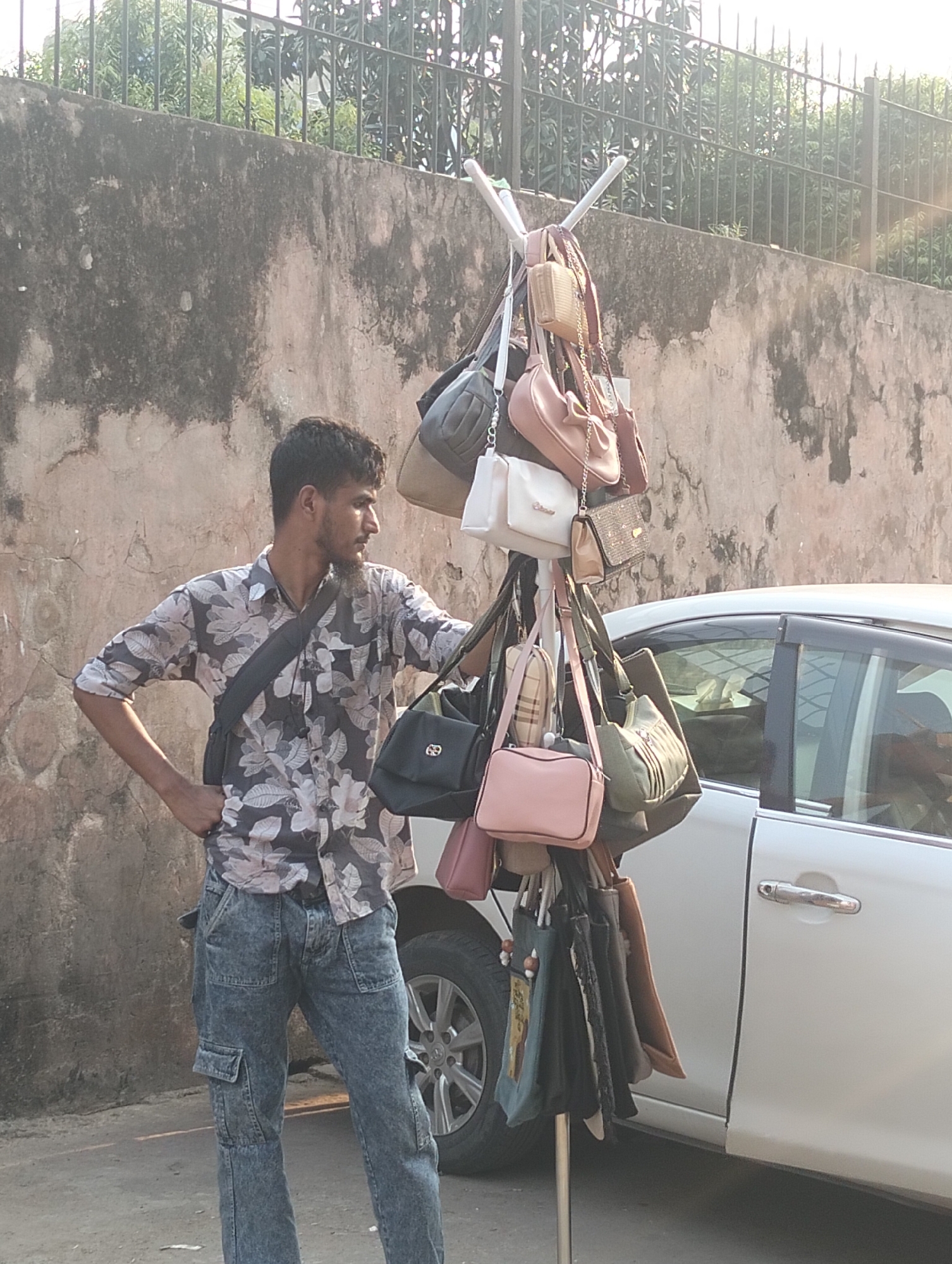 An outdoor scene captures a man standing next to a makeshift rack laden with handbags. The man is light-skinned with dark hair and a beard, wearing a floral print shirt and blue jeans, and carrying a shoulder bag. He is positioned next to a vertical stand that holds an array of handbags in various colors and styles. To the right of the stand, a white car is parked. The backdrop consists of a textured wall with an iron fence visible above. The overall setting suggests an urban environment, possibly a street in Dhaka, Bangladesh.