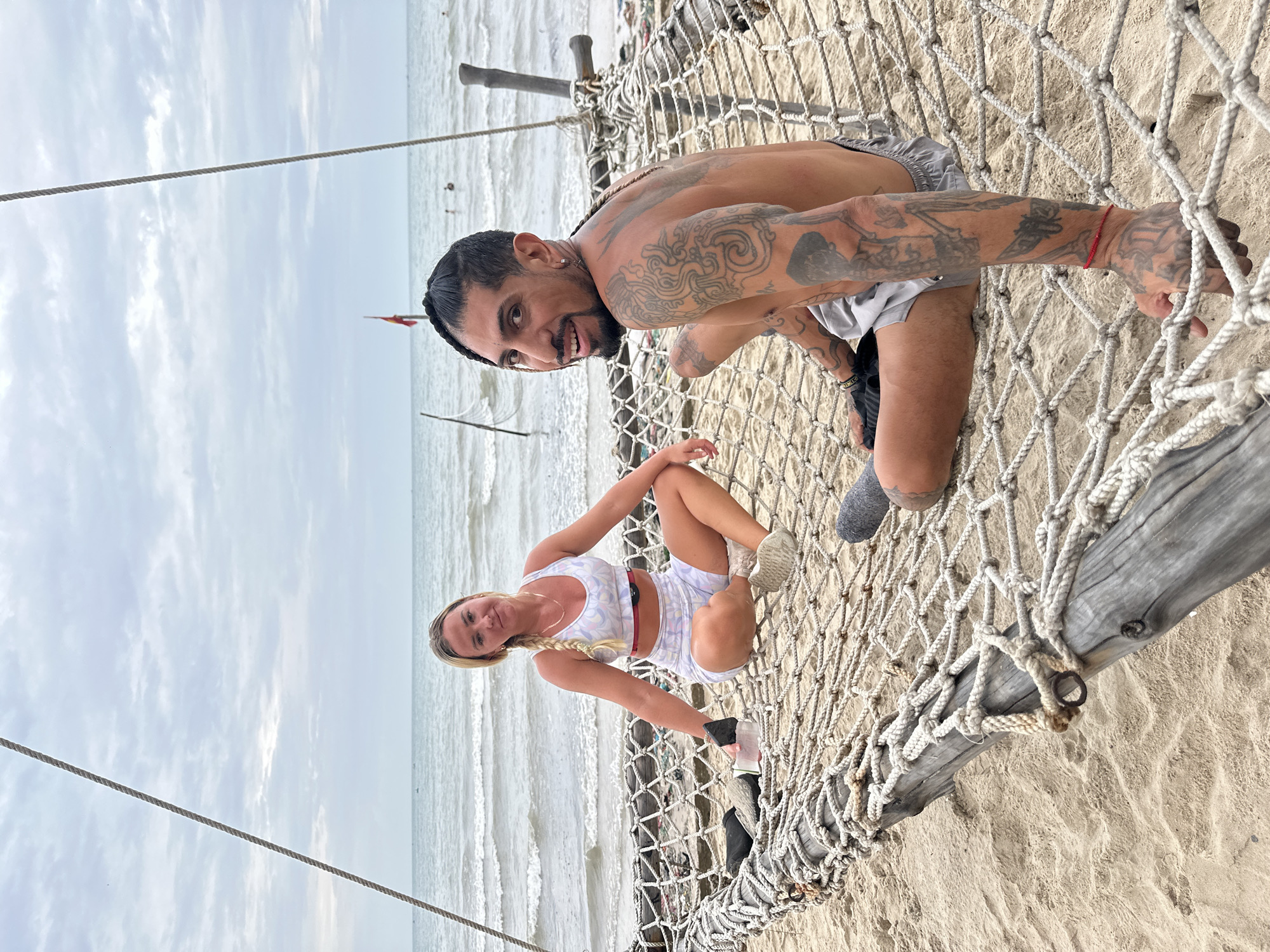 Two individuals are seated on a large, rustic net hammock situated on a sandy beach. The hammock is constructed from thick, coarse rope and supported by weathered wooden beams. The beach is composed of light-colored sand. In the background, the ocean stretches to the horizon under a cloudy sky. The water has moderate waves. A beach volleyball net and some debris are visible near the water's edge.
The individual on the left is a woman with blonde hair styled in a braid. She is wearing a light-colored athletic top and matching shorts with a floral pattern. She is seated with her legs crossed.
The individual on the right is a man with dark hair styled in braids and a beard. He is shirtless, revealing extensive tattoos on his arms and torso. He is wearing light-colored shorts. He is looking towards the camera and smiling.
The scene appears to be located on a beach in Mũi Né, Vietnam, a popular coastal resort town known for its beaches and sand dunes. The presence of the net hammock and the ocean backdrop are consistent with the typical landscape of Mũi Né.