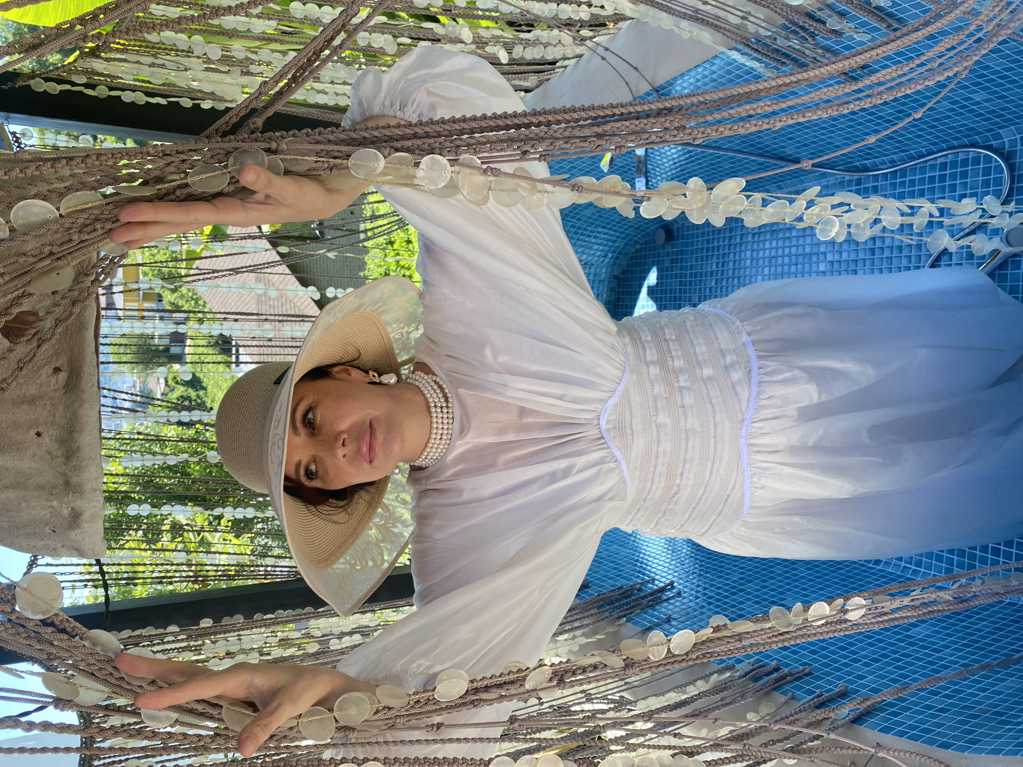 A woman wearing a white dress, a wide-brimmed hat, and pearl necklaces poses in what appears to be an outdoor shower area with blue tiled walls and a showerhead visible. She is surrounded by hanging decorations made of beads and shells. In the background, greenery and buildings are visible, suggesting a residential area in a city like Sochi, Russia. The woman is holding onto the hanging decorations with both hands.