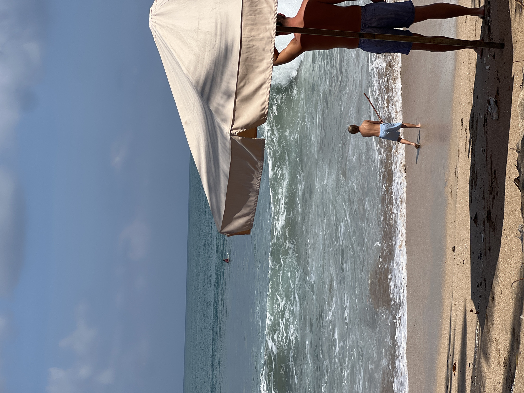 The image depicts a beach scene in Kuta, Indonesia. In the foreground, a person is standing under a large, beige beach umbrella, holding onto its pole. To the left of this person and further back towards the water, a child is standing on the wet sand and appears to be holding a stick or a toy. The child is facing the ocean, where waves are breaking. In the distance, a lone surfer is visible in the ocean. The sky above is blue with scattered clouds. The sand on the beach is light brown and shows signs of debris.