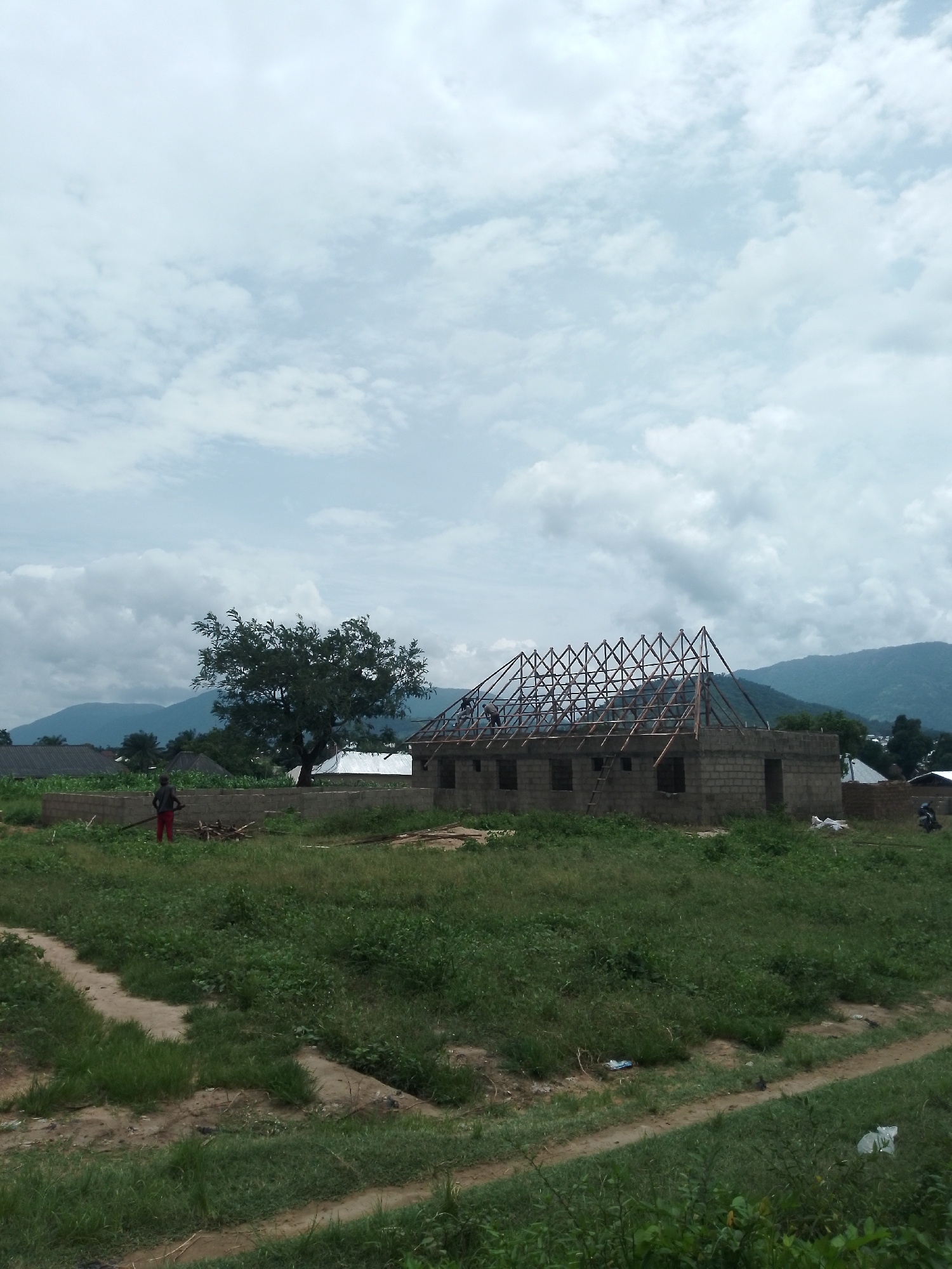 A vertical medium shot captures an outdoor scene of a house under construction in a rural or suburban environment. The sky is overcast with scattered clouds. In the foreground, a grassy, uneven terrain is visible with a dirt path. In the middle ground, a partially built house constructed of concrete blocks is the central focus. The roof framing, made of wooden beams, is in place, and several people are working on it. A large tree stands to the left of the house. Further in the background, other buildings with various roofing materials, including some with corrugated metal and some with grey, sloped roofs, are visible, suggesting a small settlement. Distant, hazy blue mountains form the backdrop. A person in red pants and a dark shirt stands near a low brick wall in the grassy area to the left of the construction site. Various construction materials and debris are scattered around the area.