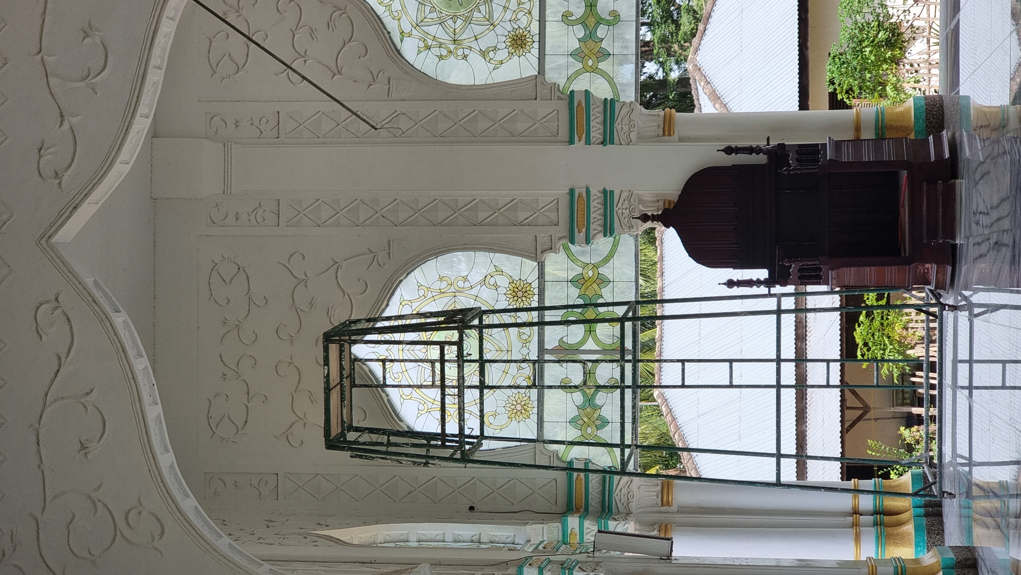 The image depicts the interior of what appears to be a mosque in Lapang Timu, Indonesia. The foreground is dominated by a polished, reflective floor. In the center, a tall, dark wooden pulpit with ornate carvings is visible. To the left of the pulpit, a green metal scaffolding structure stands, possibly for maintenance or construction. Behind these elements, an arched stained-glass window with intricate floral patterns in yellow, green, and white is prominent. The window overlooks an outdoor scene with white-roofed buildings and lush greenery. The architecture features white walls adorned with relief carvings and decorative columns with gold and turquoise accents. The overall impression is one of a sacred space with elements of both traditional and decorative design.