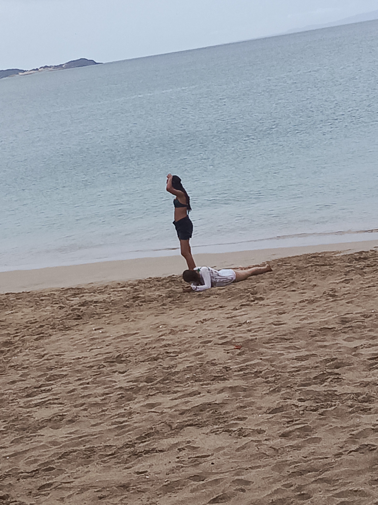 Two individuals are on a sandy beach by the sea, with a distant landmass visible on the horizon. In the foreground, one person is lying prone on the sand, facing towards the sea and wearing a white shirt and tan shorts. A second person stands upright, facing away from the sea, with their hands raised above their head, wearing a dark bikini top and dark shorts. The second person is positioned on the back of the first person. The sea is calm and a pale blue-grey, with a clear distinction between the water and the sandy shore. The sky is overcast and a pale grey. This scene appears to be in Juan Griego, Venezuela, given the characteristic landscape of the island of Margarita.