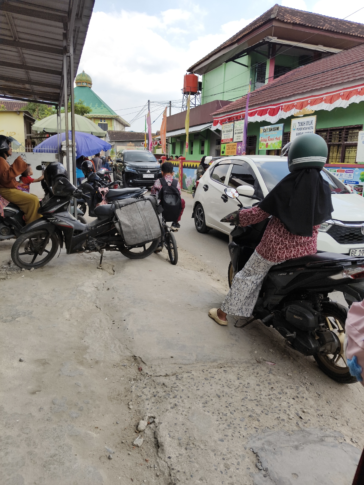 The image depicts a street scene in Bandar Lampung, Indonesia. In the foreground, a woman wearing a black hijab and a patterned long-sleeved shirt and patterned pants rides a black motorcycle. She is wearing a green helmet and beige slip-on shoes. The motorcycle has a large grey bag attached to the back. To her left, another black motorcycle is parked. A person wearing an orange hijab and a child are seated on this motorcycle. Behind the motorcycles, a black Mitsubishi Pajero Sport SUV is visible. Further in the background, a green building with a dome, possibly a mosque, is seen. A light green school building with a red and white banner is also present. The street is paved with concrete and appears to be in a state of disrepair in some areas. There are several people and parked motorcycles, suggesting a busy area. The overall scene captures a typical day in a neighborhood with a mix of residential and commercial buildings.