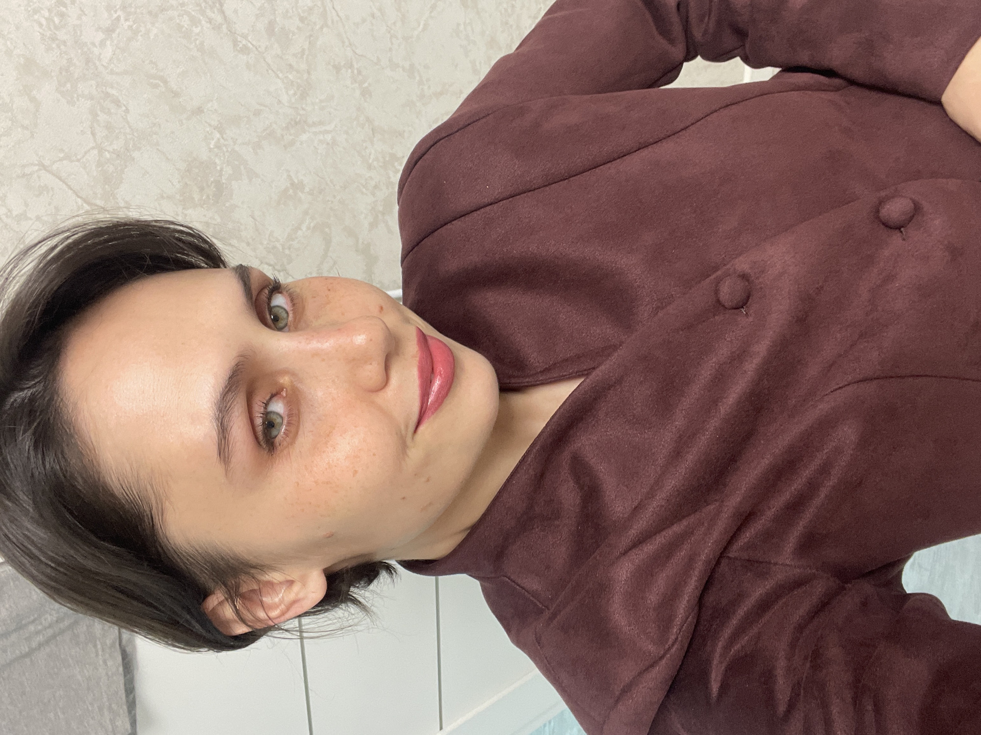 A woman with short, dark hair and light skin is facing the camera, slightly turned to her right. She has green eyes and freckles. She is wearing a brown, buttoned-up jacket. The background is a light-colored wall with a subtle pattern and a section of white wall with a hint of blue, suggesting a bathroom. The location is unknown and cannot be determined from the image. No information suggests the location is in the city of Izhevsk, Russia.
