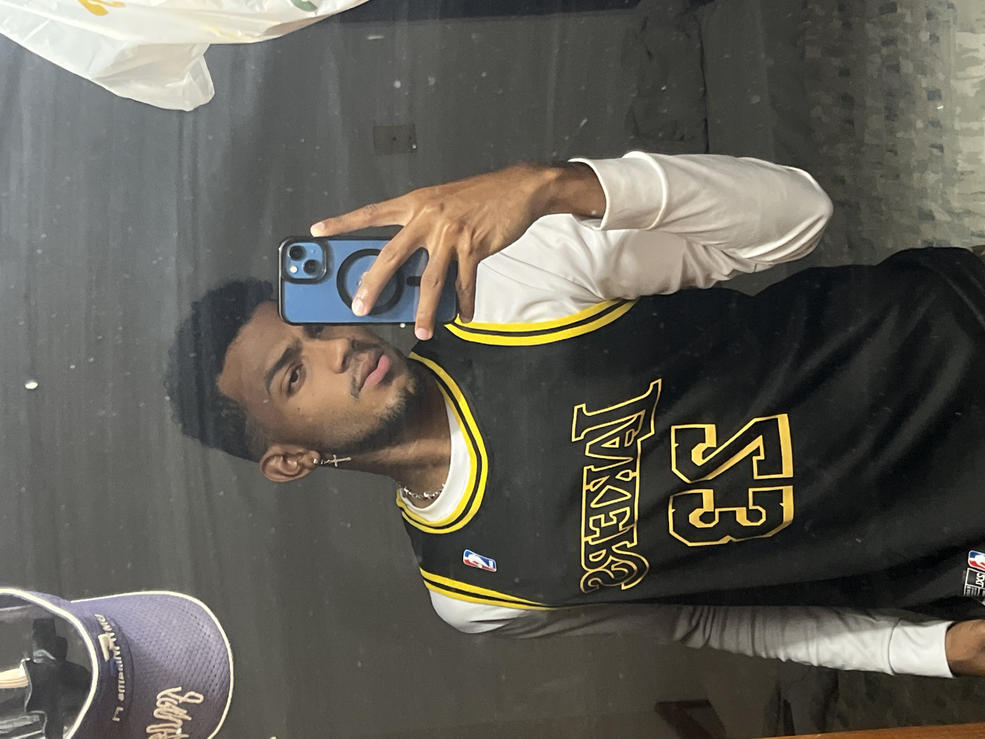 A young, dark-skinned man is taking a selfie in front of a mirror. He is wearing a black Los Angeles Lakers jersey with the number 53 in gold, a white long-sleeved shirt underneath the jersey, and a silver chain necklace. He is holding a blue iPhone in his right hand. The reflection in the mirror shows a Subway bag in the top left, a baseball cap in the bottom left, and a gray wall behind the man. The location is unknown.