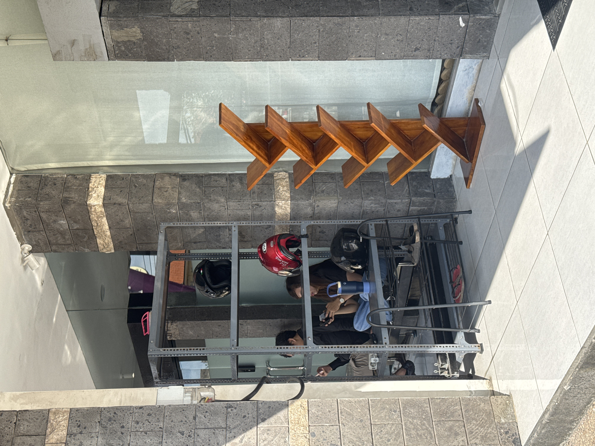 The image shows a street scene in Kuta Selatan, Indonesia. Two people are examining items within a metal shelving unit. The unit holds three motorcycle helmets (one red, two black), among other items. Above the shelving unit is a storefront window, partially covered by a retractable awning. Above the awning are five wooden triangular shelves attached to the building's facade. The sidewalk is composed of light grey tiles. The overall scene appears to be outdoors, likely a shop or business along a pedestrian area.