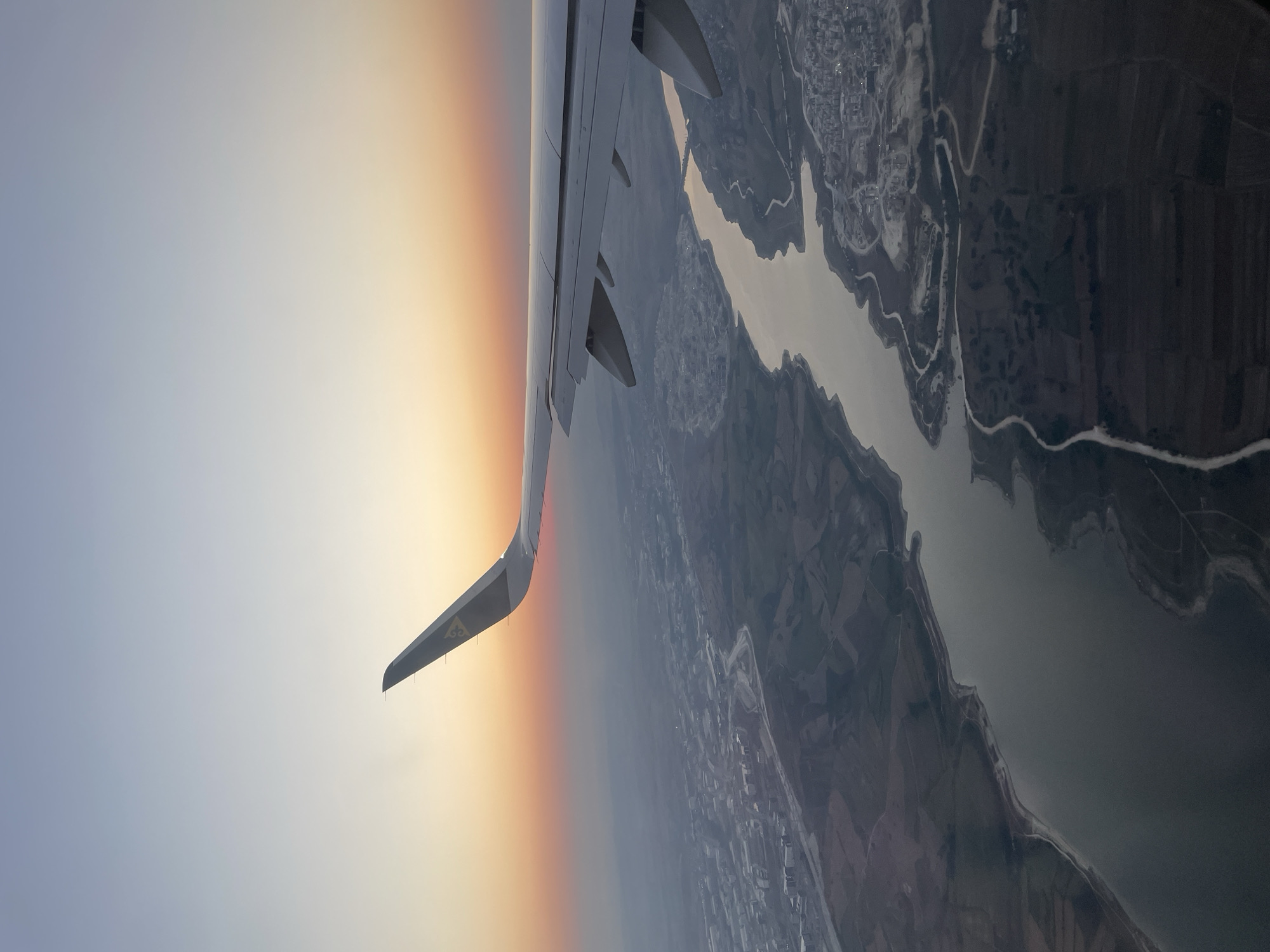 An aerial view from an airplane window shows a serene landscape at dusk. A prominent airplane wing, painted white with a yellow logo on its tip, extends from the right side of the frame, diagonally across the image. Below the wing, a body of water, likely a river or a reservoir, meanders through the terrain. The water reflects the soft, warm hues of the setting sun, creating a shimmering effect.
The land surrounding the water is a patchwork of cultivated fields and a densely populated urban area. Buildings are visible in clusters, interspersed with roads that wind through the city. The cityscape appears somewhat hazy, characteristic of a twilight scene. The horizon is marked by a gradient of colors, transitioning from a soft orange glow near the horizon to a pale blue sky above. The location is identified as Beykoz, Turkey. No people or specific actions are discernible in this static aerial scene.