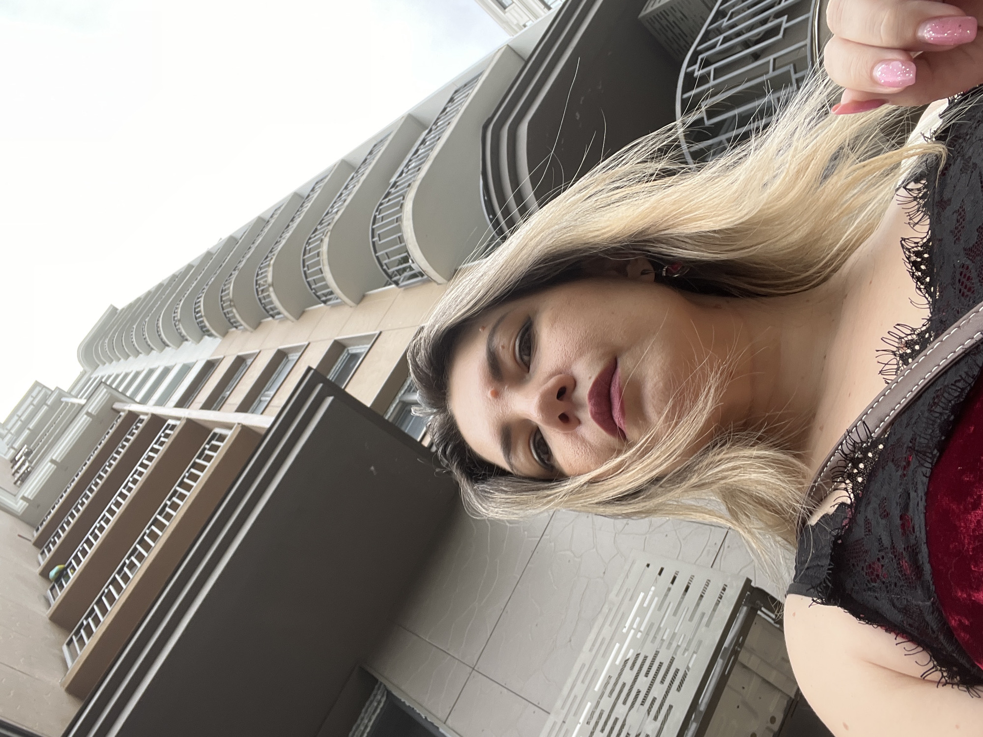 The media file is a selfie taken by a woman with blonde hair and dark lipstick, standing outside a modern apartment building with many balconies. The building appears to be tall, with a light-colored facade and dark window frames. The sky is overcast, suggesting it might be a cloudy day. The woman is wearing a dark, lacy top. The image is taken from a low angle, looking up at the building and the woman. The overall impression is a candid shot taken during a casual outing in an urban environment. No text is visible in the media.
