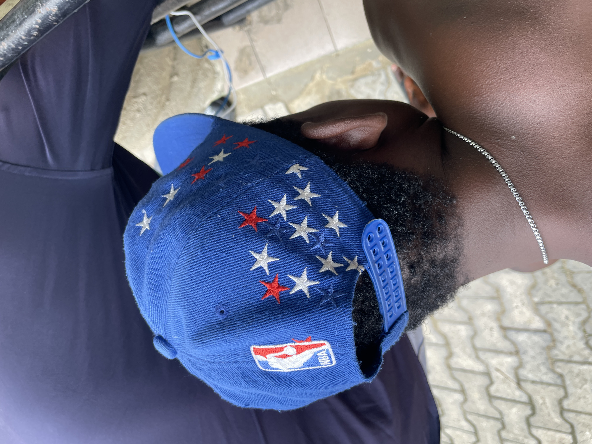 A person wearing a blue baseball cap with white and red star designs and an NBA logo is shown in the image. The person has dark skin and a beard and is wearing a silver chain necklace. The person is also wearing a dark blue shirt. The location appears to be outdoors with concrete or paved ground. There are also metal bars and wires visible in the background. The image was taken in the city of Ado, Nigeria.