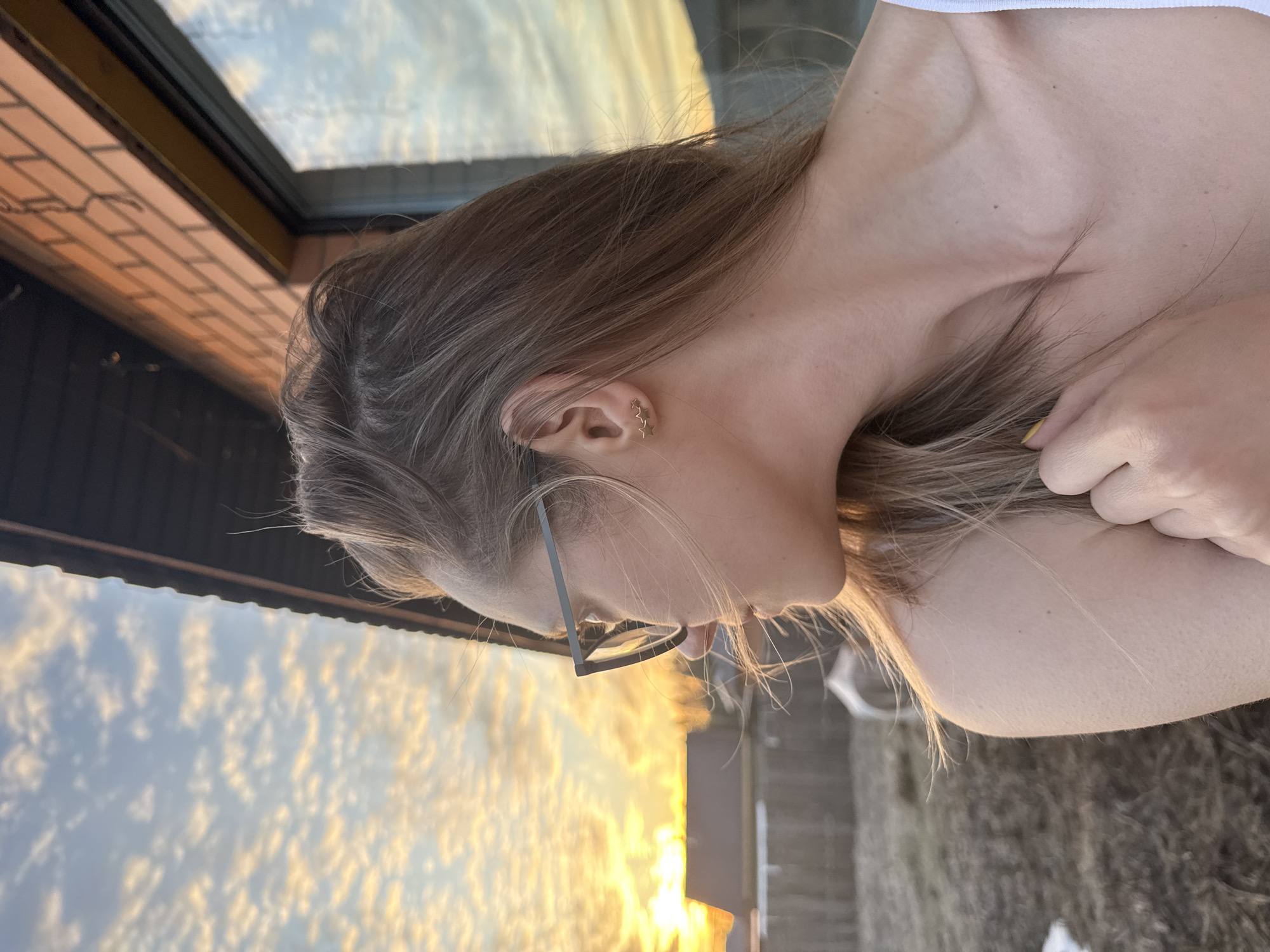 A profile shot captures a person with light skin, donned in glasses and wearing star earrings, with a glimpse of a light-colored, possibly white, garment near their shoulder. Their focus seems to be directed downwards, while the backdrop showcases a cityscape bathed in the warm hues of a sunset. The building's facade exhibits brickwork near a window, and the sky is painted with the scattered clouds of twilight.
This picture was taken in Chaikovsky, Russia.