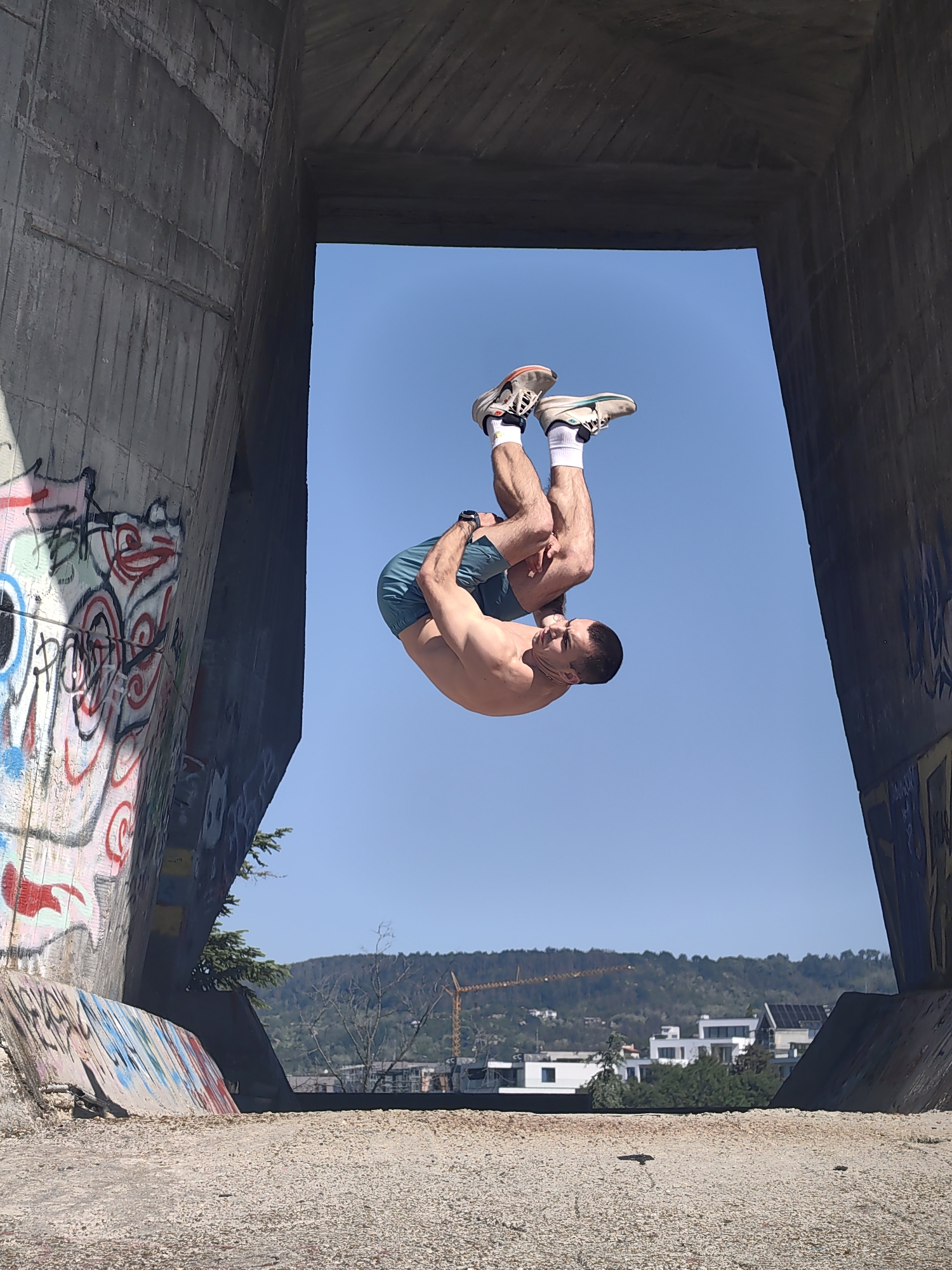 A man is captured mid-air performing a backflip, framed by a concrete structure covered in graffiti. The man is wearing blue shorts, white socks, and running shoes. His torso is bare. The backdrop reveals a cityscape with buildings and a hill. A construction crane is visible in the distance. The scene is located in Varna, Bulgaria, within one of the city's urban parkour locations near a bridge or overpass, recognizable by the specific style of concrete and graffiti found in such areas. The ground is covered in light-colored gravel or concrete rubble. The sky is clear and blue.