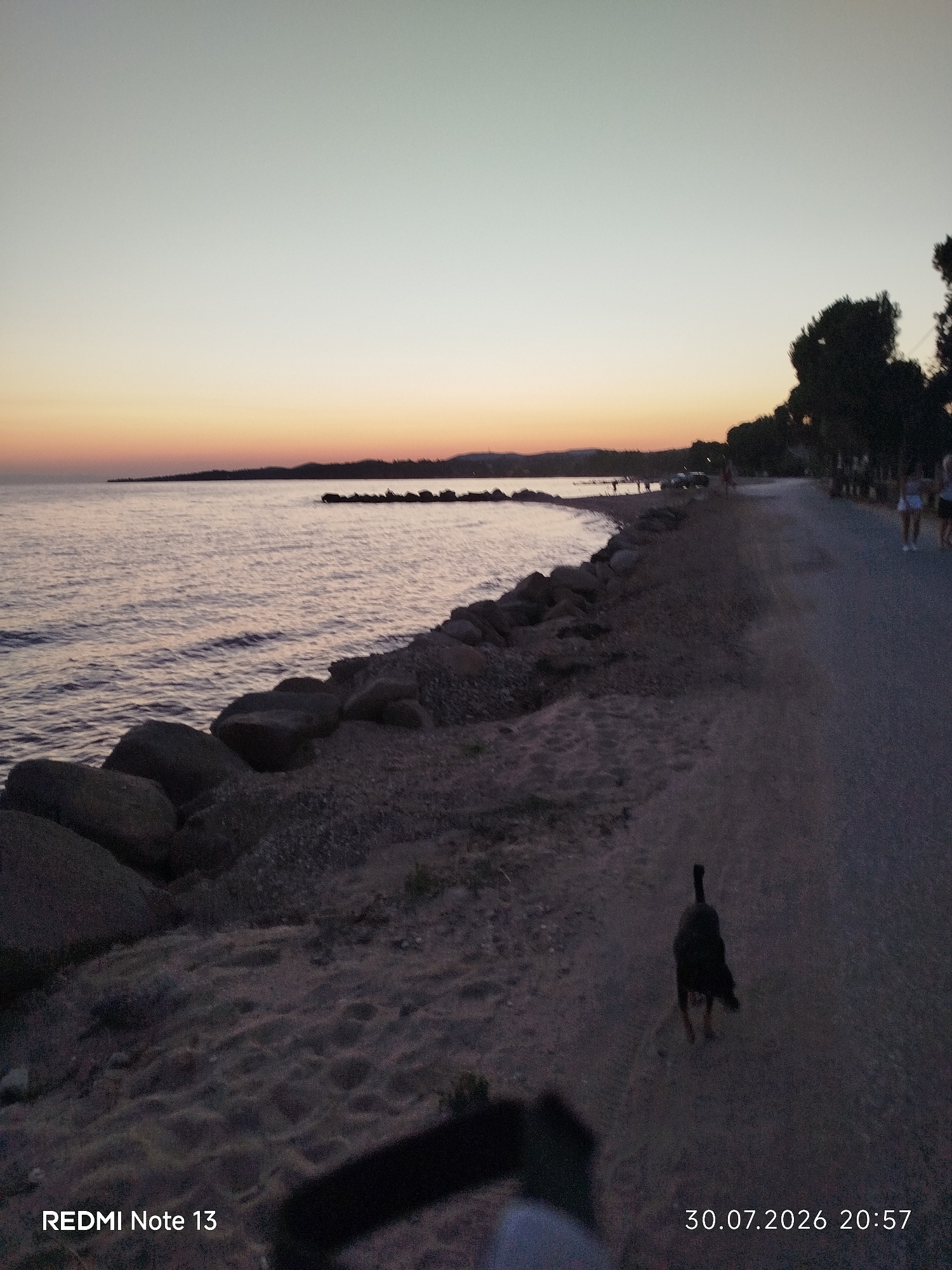 An outdoor, medium-wide shot captures a coastal scene at dusk in Sithonia Municipal Unit, Greece. The horizon features a tranquil sea meeting a gradient sky transitioning from pale yellow to light grey. A shoreline of dark rocks leads to a sandy beach. On the right, a paved walkway borders the beach, lined with silhouetted trees. A solitary dog, dark in color, walks away from the camera on the walkway. In the distance on the walkway, a few indistinct figures and vehicles are visible. The bottom left corner displays "REDMI Note 13" and the bottom right corner shows "30.07.2026 20:57".