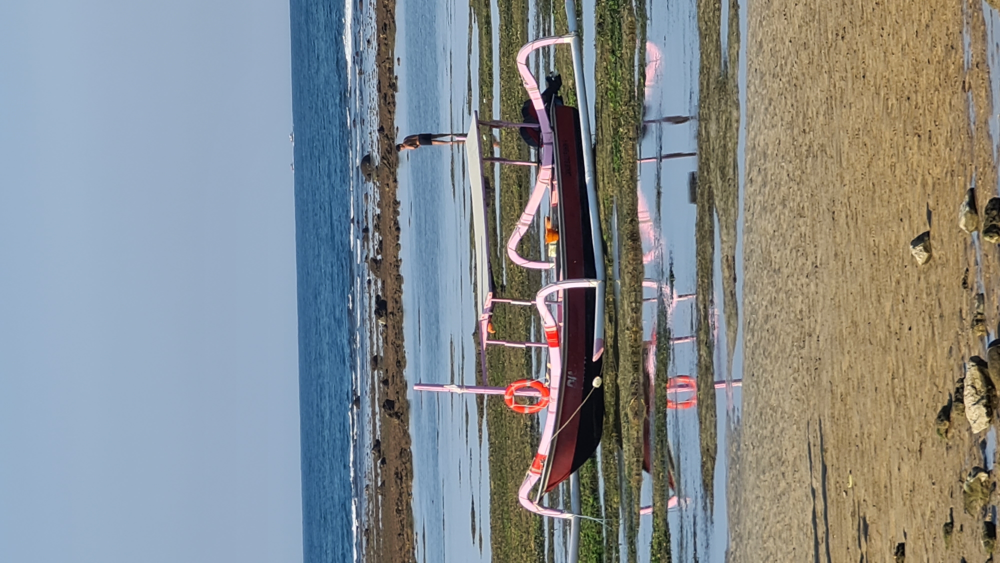A traditional outrigger boat, painted dark red with bright pink accents, rests on a shallow, algae-covered seabed. The boat features a central canopy supported by pink poles and a life preserver hanging on its side. A lone individual, wearing dark shorts, stands in the water behind the boat, looking away from the camera. The scene is set at low tide, with shallow pools of water reflecting the boat and the sky. In the distance, the blue ocean meets the horizon under a clear sky. This image was taken in Denpasar, Indonesia.