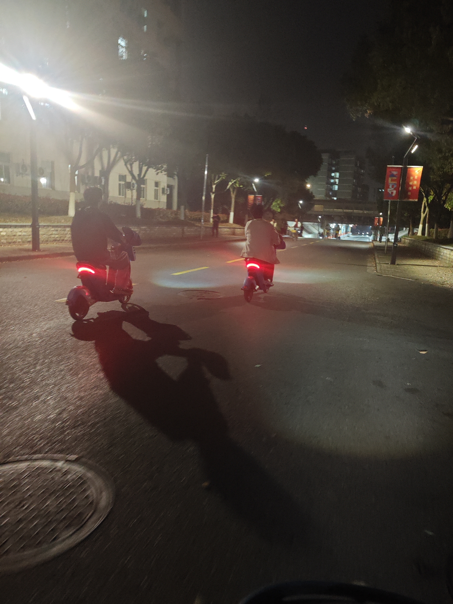 Two people are riding electric scooters on a road at night in Jiangning District, China. The road is paved and lit by streetlights. One person is wearing dark clothing, and the other is wearing light clothing. The scooters have red taillights. There are buildings and trees in the background. There are two banners on a pole in the background, to the right.