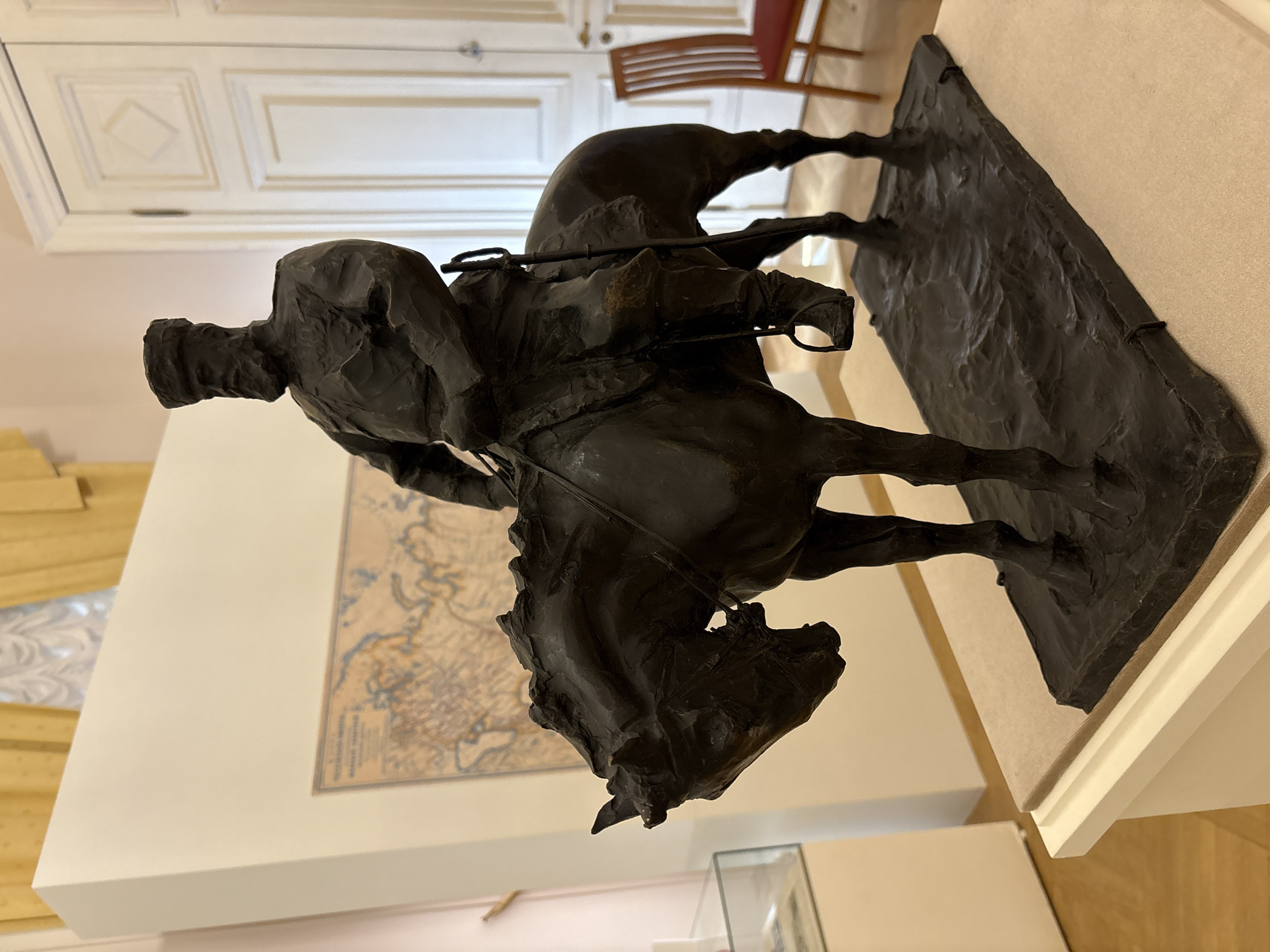 The image shows a bronze sculpture of a Cossack on horseback, positioned on a cream-colored plinth within a museum setting. The sculpture depicts a rider wearing a cap and a dark, heavily draped coat. The horse is shown mid-stride, with its forelegs extended. The backdrop includes a light-colored wall displaying a framed map of a region with rivers and towns. In the background, a section of a light-colored door with decorative panels is visible, along with a wooden chair. The setting suggests an interior space in the city of Saint Petersburg, Russia.