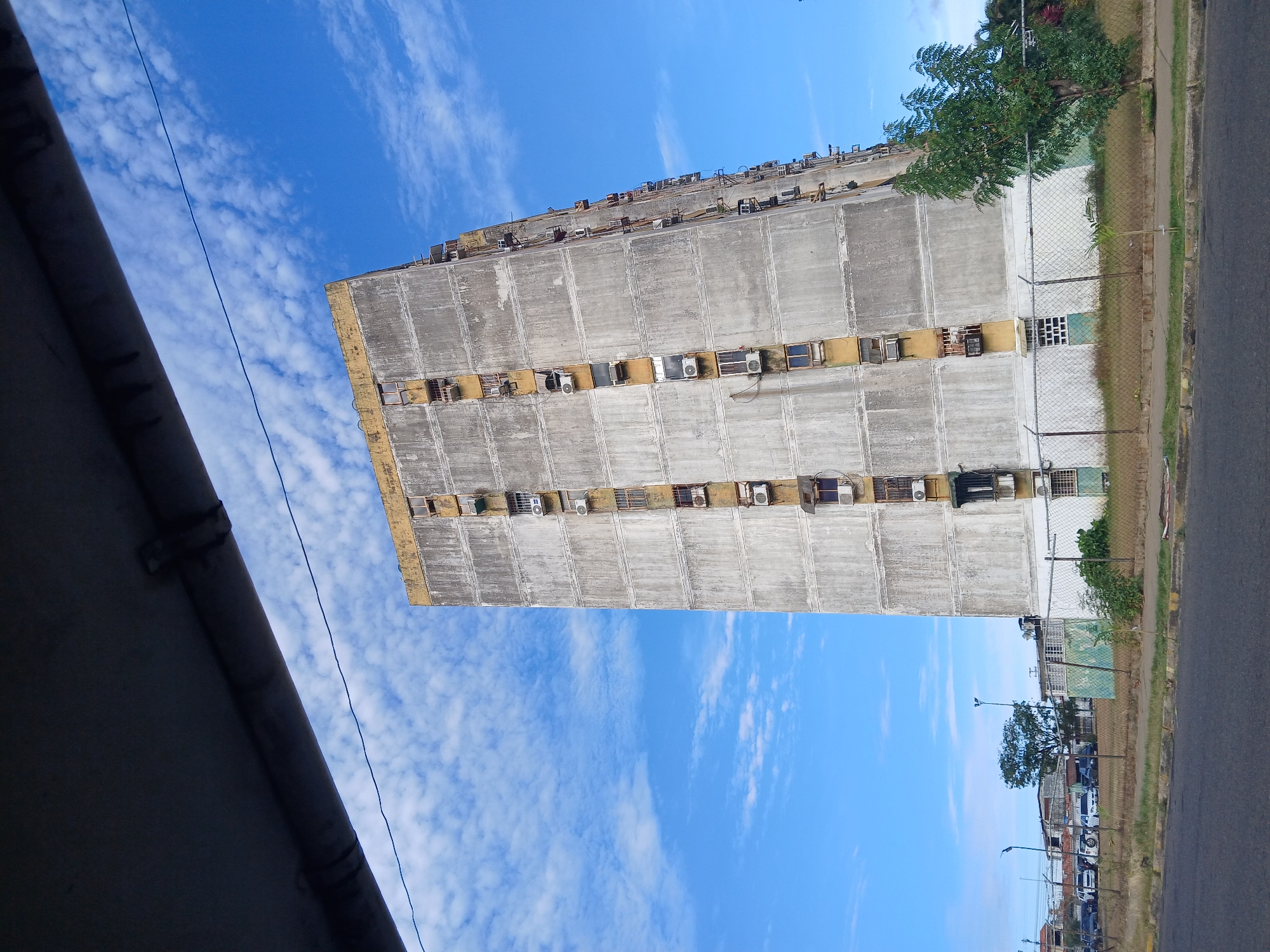 The image depicts a tall, dilapidated concrete building, likely an apartment block, in Ciudad Guayana, Venezuela. The building has numerous air conditioning units protruding from the windows on each floor. The concrete facade shows signs of weathering and staining. A section of a corrugated metal roof is visible in the lower left corner of the frame, suggesting the vantage point is from a nearby structure. A chain-link fence separates the building from the street and sidewalk. The sky is blue with scattered clouds. A street runs alongside the building with parked cars and trees visible in the background.