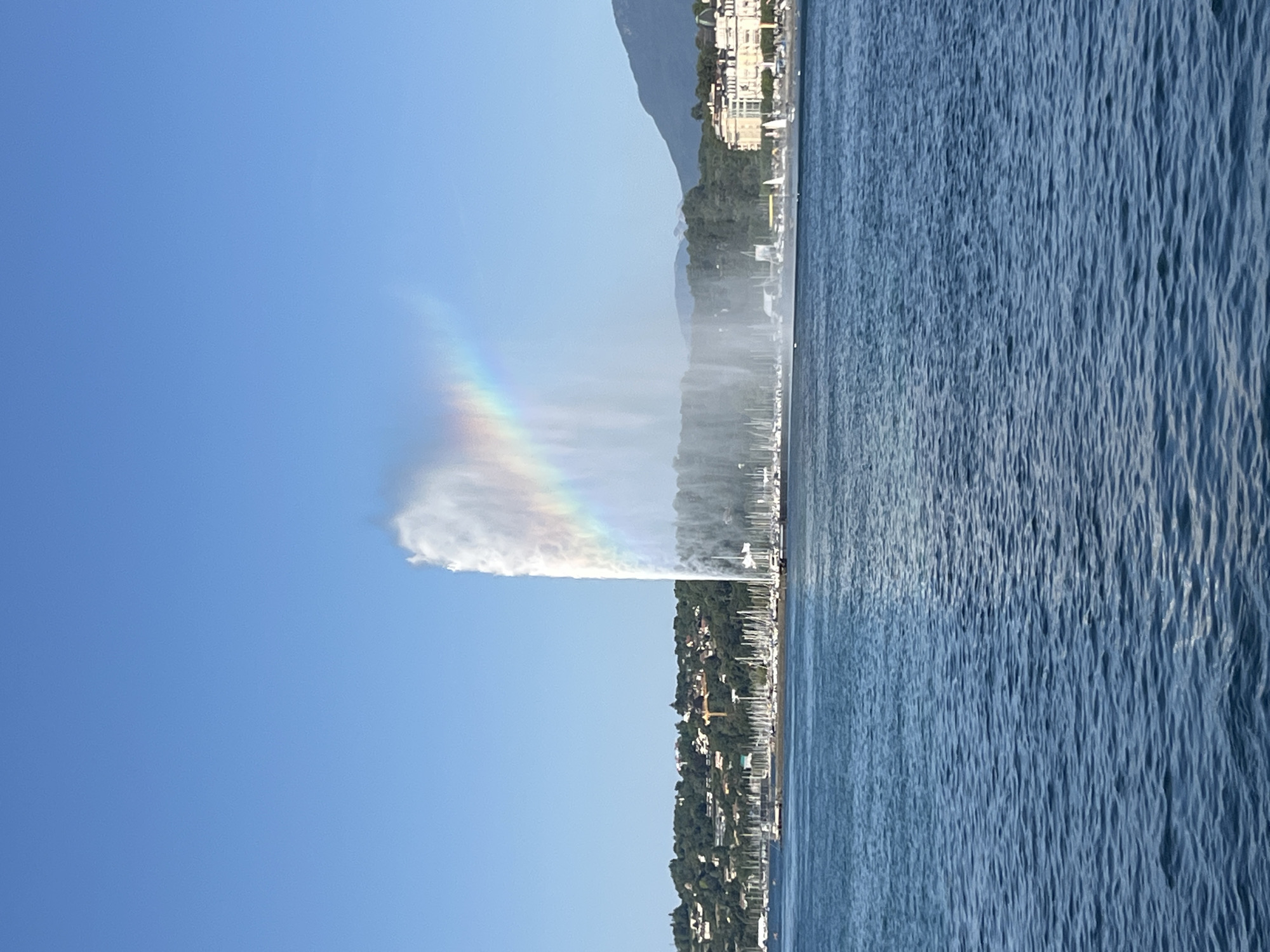 This is a photograph of the Jet d'Eau fountain in Geneva, Switzerland, taken from the water. The fountain, a tall plume of water reaching high into the clear blue sky, is the central focus. A rainbow is visible within the mist of the fountain, arcing across its upper section. The water is choppy and blue, with a few ripples and reflections. In the background, a shoreline with trees, buildings, and a large number of sailboats docked in a harbor can be seen. Mountains are visible in the far distance. The sky is a solid, cloudless blue. There are no people or animals readily visible in the image.
