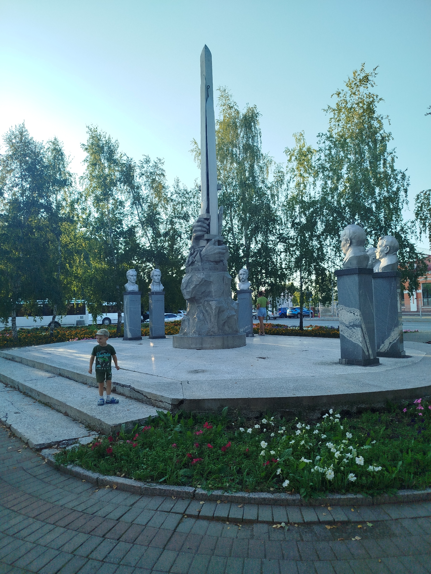 A young boy wearing a green t-shirt and shorts stands in front of a monument in Yalutorovsk, Russia. The monument features several busts of men on pedestals, as well as a tall obelisk. Trees and flowers surround the monument, and a street with cars is visible in the background. The sky is clear and blue.