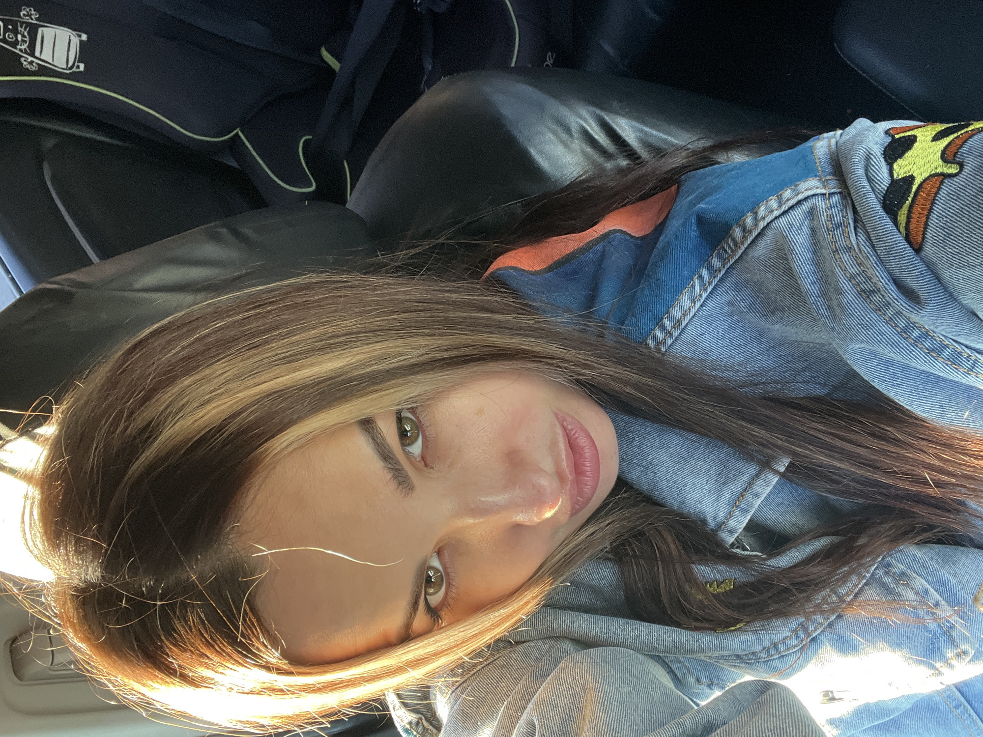 A close-up, slightly angled shot shows a person with brown hair and light highlights wearing a denim jacket. The person is indoors, possibly in a car, with a black seat visible in the background. The location is unknown.