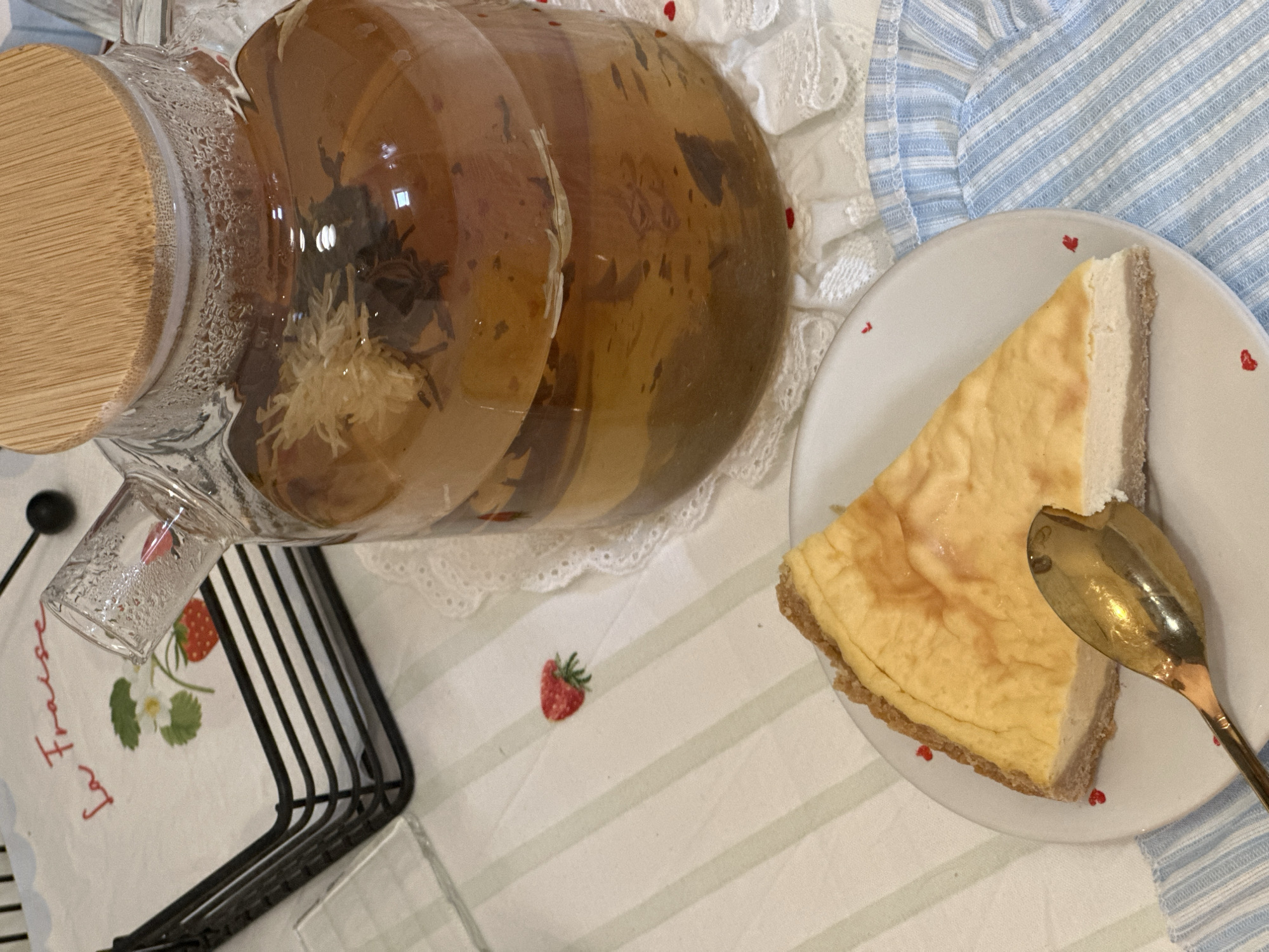 The image presents a still life of food and beverages, likely indoors. A clear glass teapot filled with tea (possibly herbal, given the flower visible inside) with a wooden lid sits on a white lacy doily atop a tablecloth with green stripes and scattered red strawberry motifs. To the right, a plate decorated with small red hearts holds a slice of cheesecake, with a golden spoon partially submerged in it. A black metal rack is positioned on the left side of the frame, partially obscured. The environment includes blue and white striped fabric, adding to the overall aesthetic. The image suggests a leisurely, potentially domestic setting. There is no information available in the image to suggest this photograph was taken in Šušanj, Montenegro.