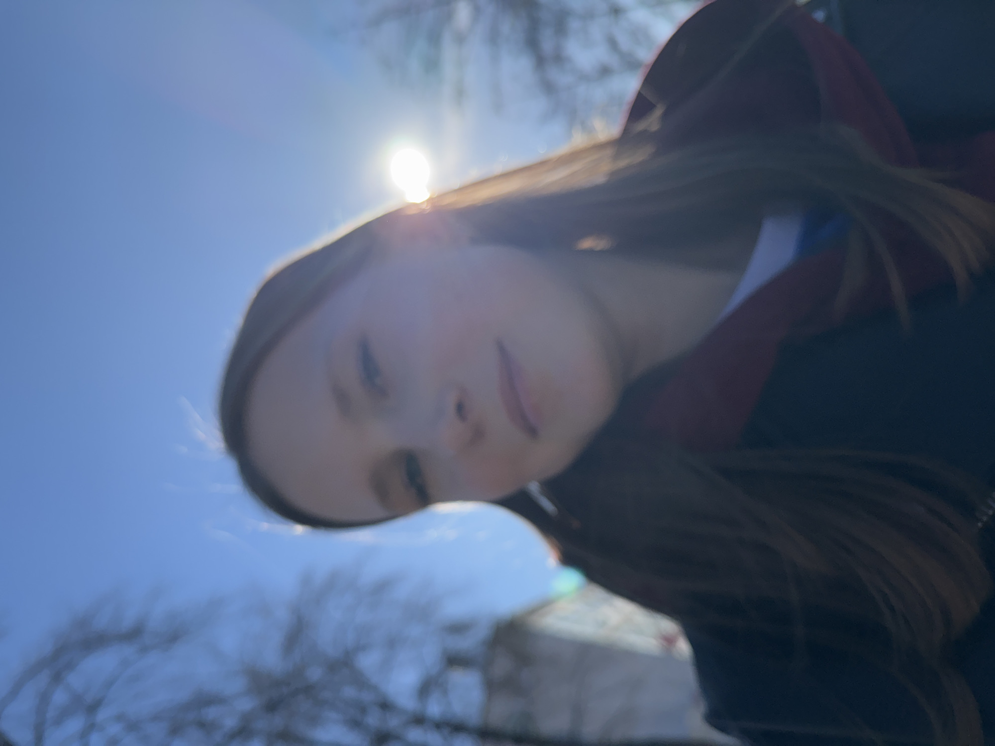 A close-up image shows a young person, likely female, taking a selfie outdoors on a bright day in Chaikovsky, Russia. The person has long, light brown hair and appears to be wearing a red hoodie or jacket over a darker top with a visible white or light blue stripe. Their face is angled downwards towards the camera, with a neutral or slightly smiling expression, though details are softened by the intense backlighting.
The sun is positioned directly behind the person's head, creating a strong lens flare and a brilliant halo effect around their hair and upper body. The sky is a clear, bright blue, indicating good weather. In the blurred background, bare tree branches are visible, suggesting the season is either late autumn, winter, or early spring. The strong sun suggests it is daytime, possibly mid-morning or afternoon. The overall composition uses a tilted angle, giving the image a casual and dynamic feel. There is no visible text in the photograph.
