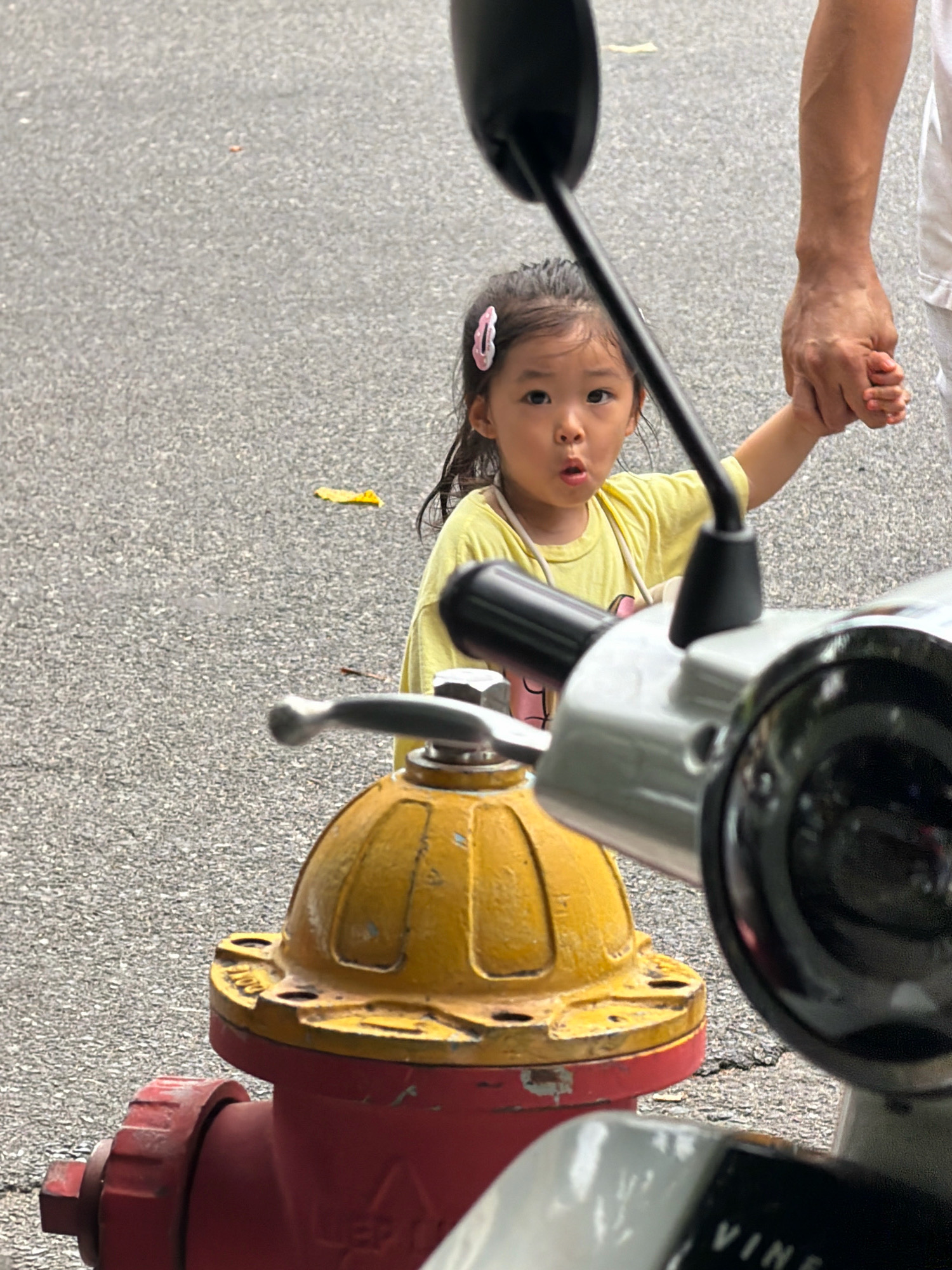 A young East Asian girl, with dark hair and a pink hair clip, stands on a paved surface, holding hands with an adult whose arm and upper torso are visible on the right. The girl wears a yellow t-shirt and has a surprised expression. In the foreground, a red and yellow fire hydrant is partially obscured by a silver and black scooter. The background is an asphalt road with some fallen leaves. The scene is likely in an urban environment, and given the context of Nha Trang, Vietnam, it is plausible this photo was taken on a street or sidewalk there.
