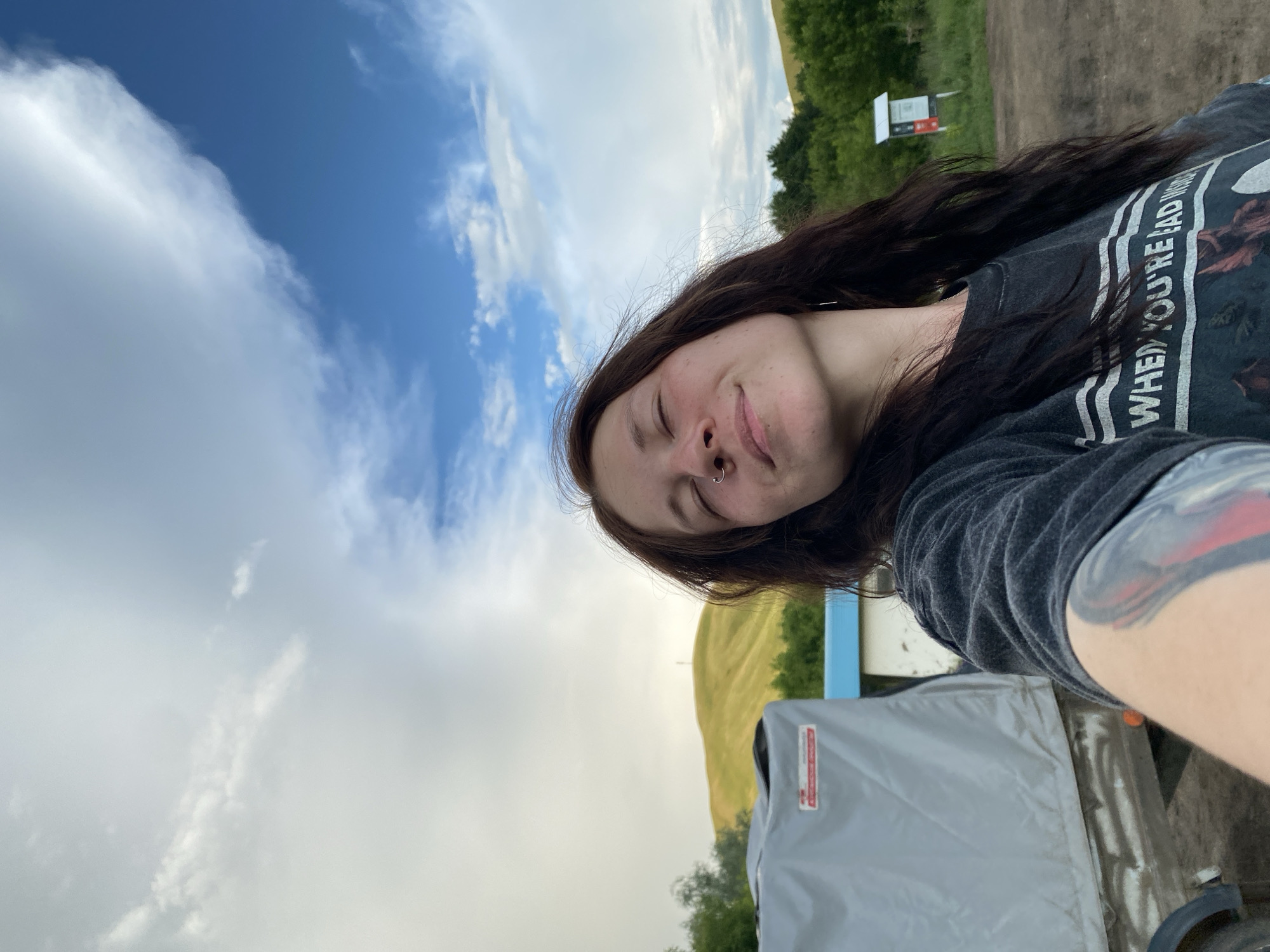 A young woman with long, dark hair and a nose ring takes a selfie outdoors. She is wearing a dark gray t-shirt with a graphic print. Her eyes are closed, and she has a relaxed smile on her face, suggesting a pleasant or contented emotion. Her arm, which is visible in the foreground, has a tattoo.
The background shows a vast sky with dramatic clouds, a mix of bright white and darker gray. Beyond the sky, there are rolling green hills and trees, indicating a rural or natural setting. A dirt road is visible in the upper right, and a signpost stands near the road, though the text on it is not legible. A light-colored tarp covers an object in the lower part of the image, with a red and white label visible on it. The overall impression is of an overcast day, possibly late afternoon, given the lighting. The scene appears to be located in Верхнеозёрное, Russia, suggesting an outdoor activity or a moment of personal reflection in a scenic location.