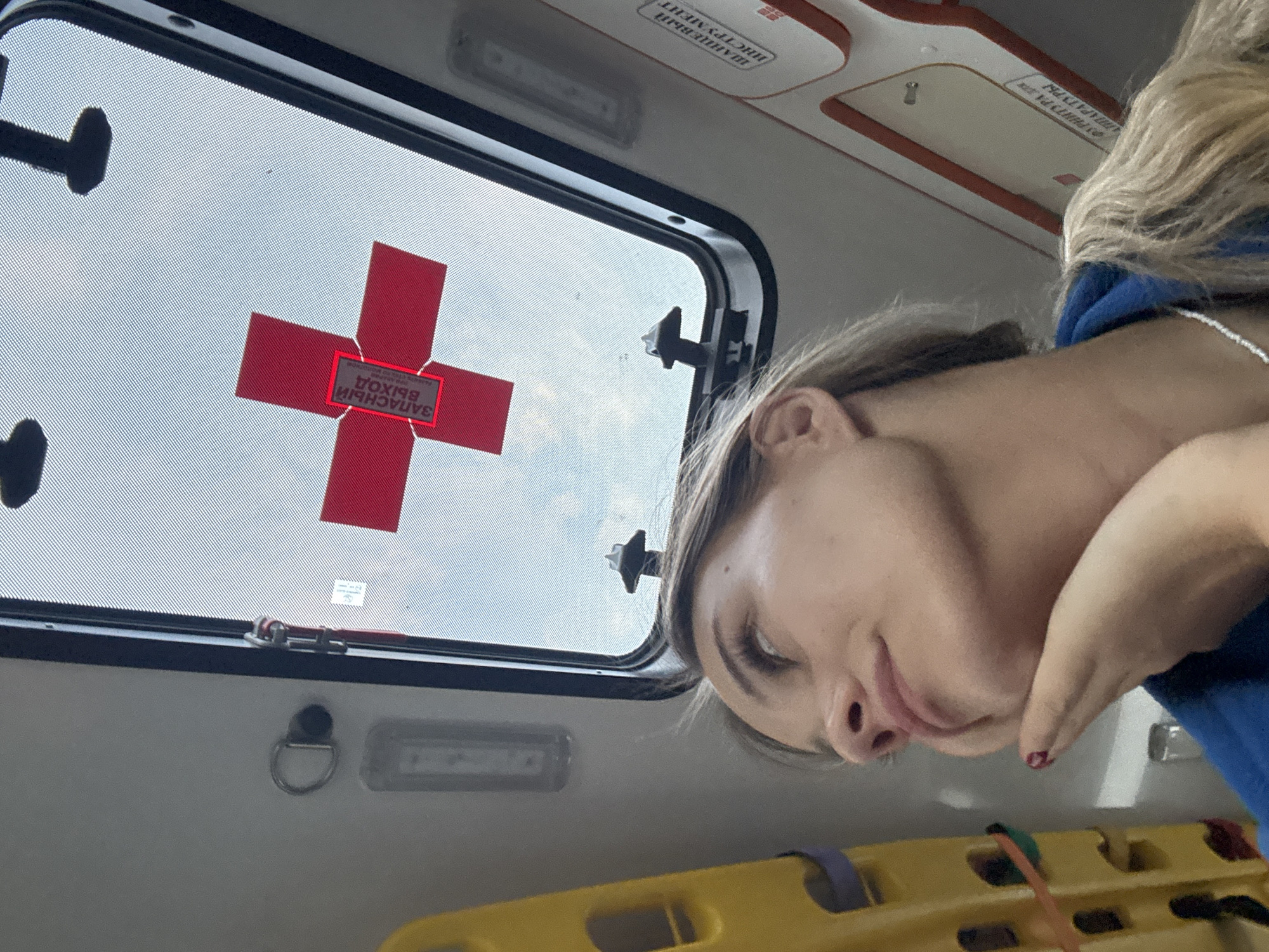 The image captures the interior of a medical transport vehicle, likely an ambulance, in the city of Chaikovsky, Russia. A young adult female is positioned in the lower left to central foreground, her gaze directed upwards and slightly to her left towards a roof window or emergency hatch. She has light-colored hair, is wearing a blue garment and a white beaded necklace. Her right hand, featuring dark red nail polish on at least one finger, supports her chin. Through the black-framed window/hatch, a bright sky is visible. A red cross symbol is affixed to the exterior surface of the window, upon which a dark grey rectangular label with white Russian text reading "ЗАПАСНЫЙ ВЫХОД" (EMERGENCY EXIT) is prominently displayed. To the left of the window frame, a silver D-ring hook is mounted on the light-colored interior wall. A portion of a yellow medical stretcher or equipment rack, including a purple strap, is visible on the far left. A long, rectangular light fixture is mounted above the stretcher. Partially visible Russian text labels on the right wall include "ШЛАНГОВЫЙ ИНСТРУМЕНТ" (Hose Tool) and "ФУРНИТУРА ДЛЯ АППАРАТУРЫ" (Fittings for Equipment).