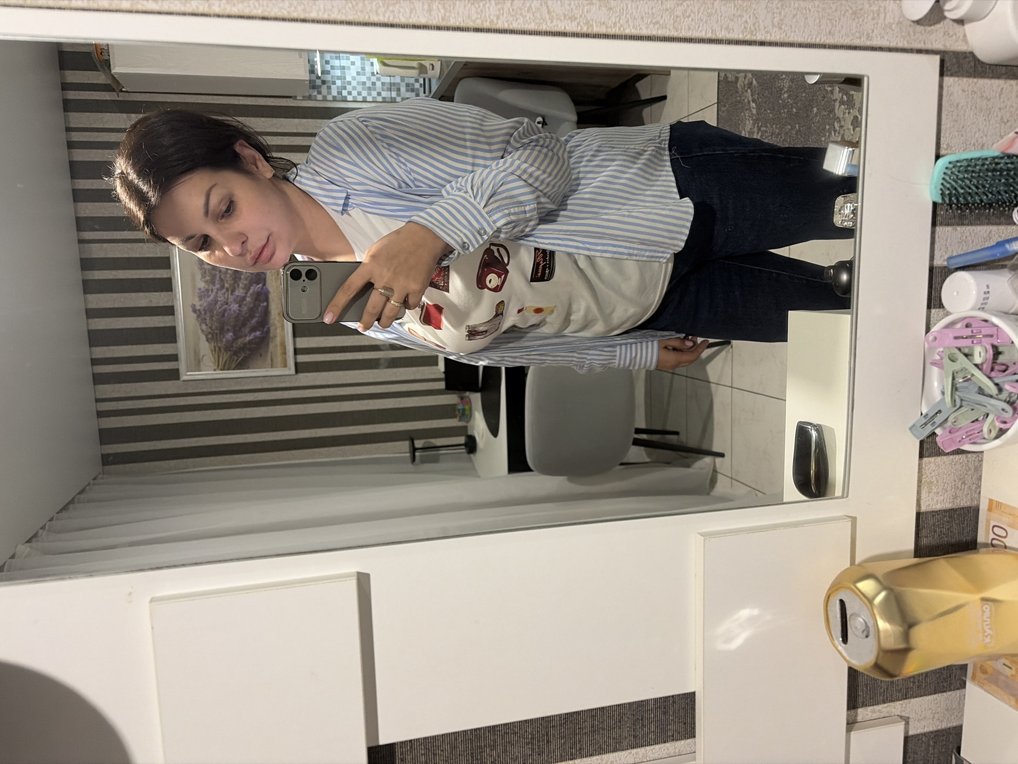 The image displays a mirror selfie of a woman taken indoors in a domestic setting, identified as the city of Sochi, Russia.
**Subject:** A woman, appearing to be in her 30s or 40s, with medium-dark brown hair pulled back, is the primary subject. She is wearing a light blue and white vertically striped long-sleeved button-down shirt, worn unbuttoned over a white t-shirt featuring multiple colorful graphic prints, including a red purse, a brown cowboy boot, a red landline telephone, and a yellow bottle. She is also wearing dark blue jeans. A gold ring is visible on the ring finger of her left hand, which is holding a silver/grey smartphone with a distinctive multi-camera module and a protective case. Her gaze is directed towards the phone screen, and her head is tilted slightly.
**Environment (Reflected in Mirror):** The background, reflected in the mirror, shows an interior space with a modern aesthetic. The wall behind her features vertically striped wallpaper with alternating dark grey/black and light grey/white bands. A framed picture depicting a bundle of purple lavender flowers is hung on this wall. To the left of the framed picture (as viewed in the reflection) is a white desk with a dark surface, a black modern-style desk lamp, and a grey office chair with dark legs. A small, colorful object rests on the desk. To the right, glimpses of a kitchen or dining area are visible, including white upper cabinets, a tiled backsplash (small, light-colored mosaic tiles), and a second grey chair. White, sheer curtains are visible hanging from a ceiling-mounted track along the far left edge of the reflected background. The floor appears to be light-colored tile.
**Foreground (Below Mirror):** A white shelf runs horizontally below the mirror. On the left side of this shelf, a gold, faceted, geometric-shaped piggy bank (or decorative container) with a coin slot is present, bearing the Cyrillic text "куплю" (buy/purchase). A partially visible 500 Ruble banknote is placed beneath it. On the right side of the shelf, a white bowl contains numerous plastic clothespins in various colors (pink, light blue, white, green). Adjacent to the bowl are a white cylindrical bottle, a blue pen, a teal/green brush, and a white pump bottle (likely a soap or lotion dispenser). Reflected on the shelf surface below the woman's torso are several objects, including a black rectangular item and a silver, reflective item.