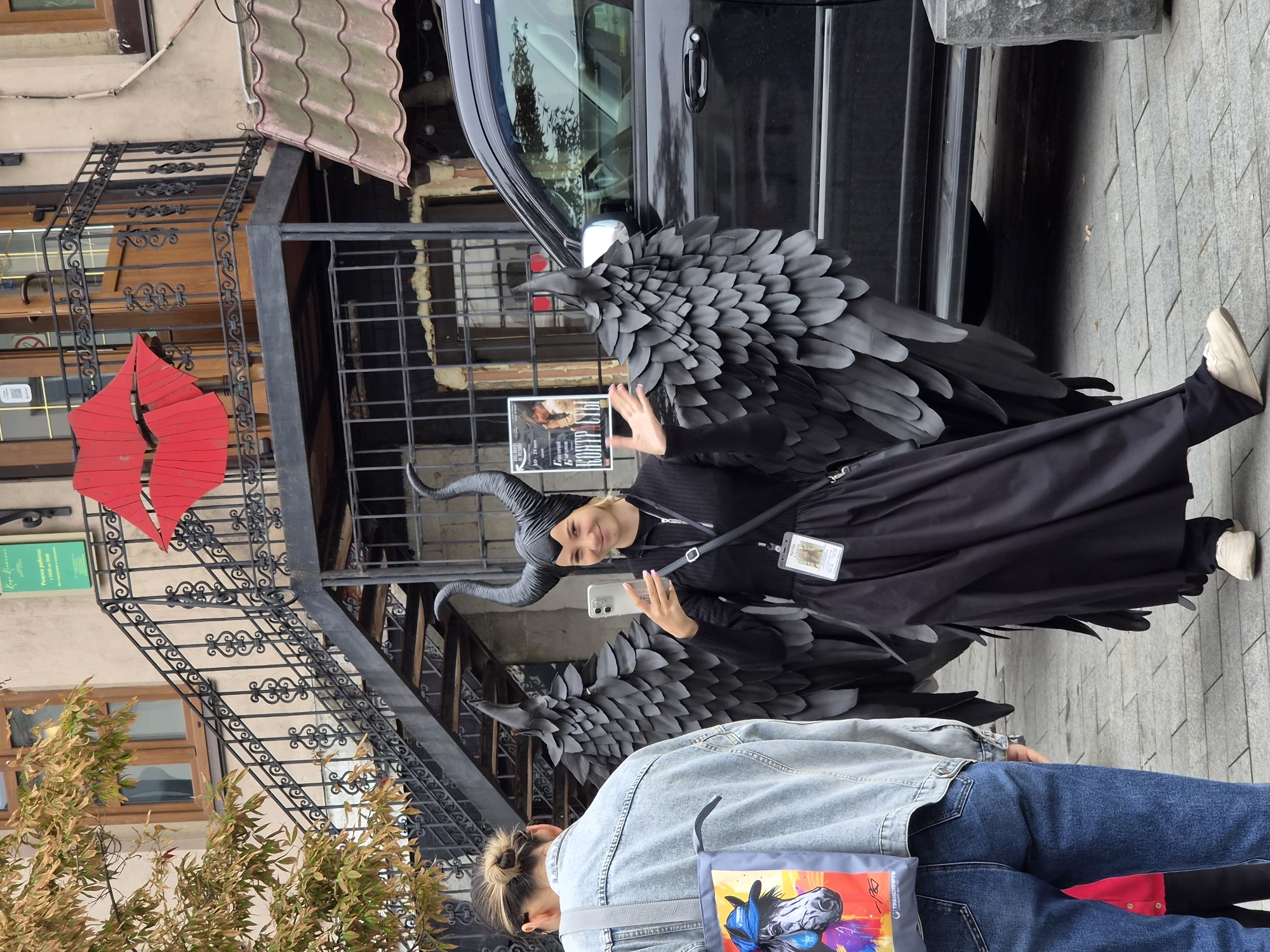 A young woman, dressed in a Maleficent costume with black horns, a black dress, and large black wings, stands in front of a black SUV. She is holding a smartphone in her right hand, taking a selfie. A woman in a denim jacket and jeans stands to her left, facing away from the camera. The background features a building with a red lip sculpture, a staircase with black railings, and a green sign with Russian text. This scene is captured on a paved street in Vladivostok, Russia.