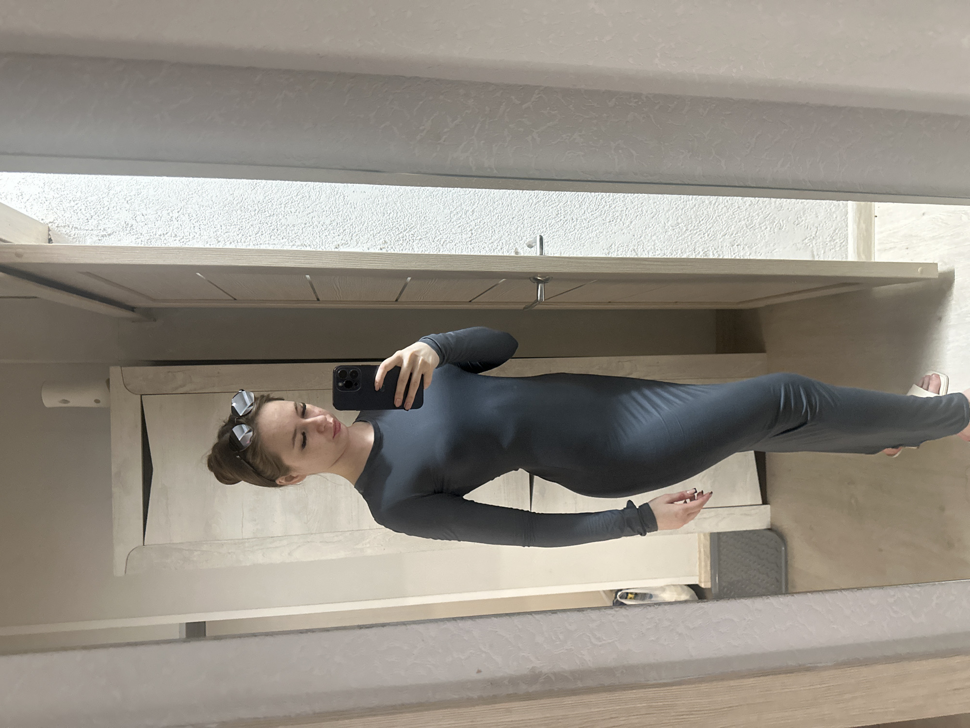A woman is taking a mirror selfie indoors. She is wearing a dark gray, form-fitting, long-sleeved dress. The room appears to be a bedroom or dressing area, containing a wardrobe or cabinet and a mirror. The location is unknown.