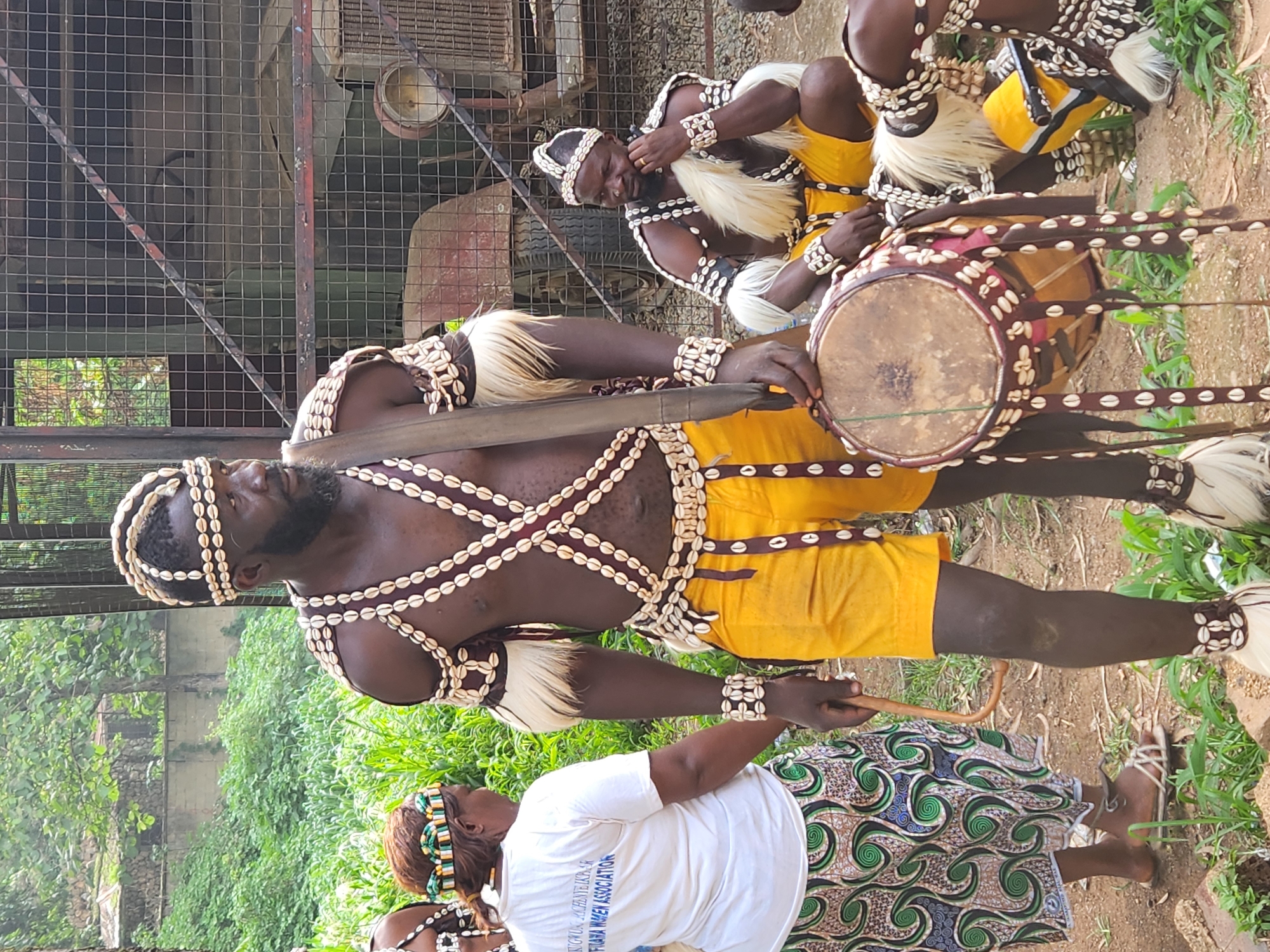 In the city of Jos, Nigeria, a group of individuals dressed in traditional attire are gathered. A man in the foreground, wearing a headdress, chest adornments, armbands, and leg bands made of cowrie shells and fabric, holds a drum. His outfit also includes yellow shorts and a sash across his chest. He has a beard and is looking to the right. To his left, a woman in a white t-shirt and a patterned skirt stands, also adorned with a beaded headband. Behind them, other individuals in similar traditional attire are visible, one of whom is seated and resting his chin on his hand. A rusty metal cage and what appears to be a vintage vehicle are in the background, surrounded by lush green foliage. The ground is a mixture of dirt and grass. The scene appears to be an outdoor cultural gathering or performance.
