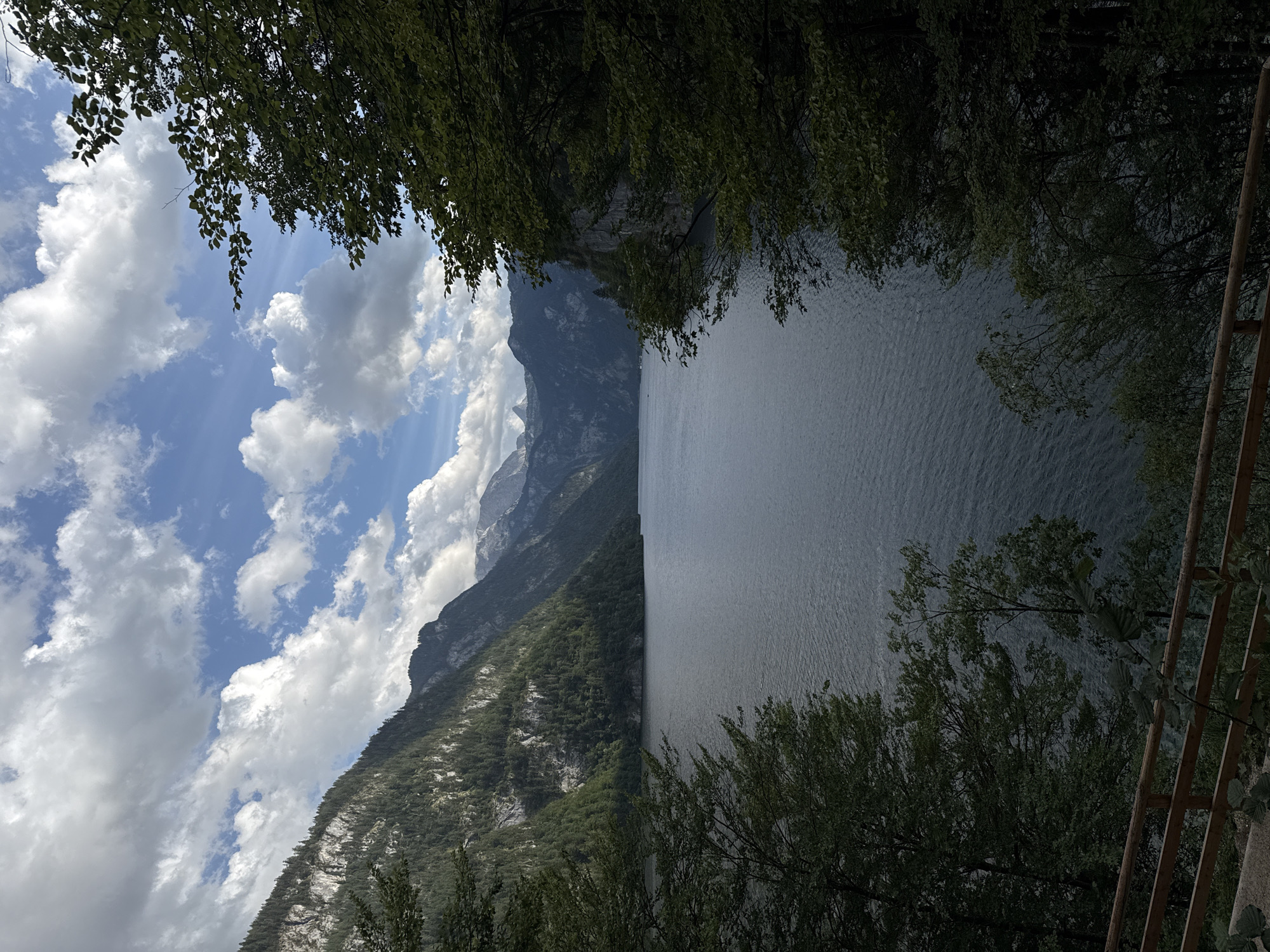 This image captures a scenic view of Königssee lake in Schönau am Königssee, Germany. The lake is framed by lush green trees in the foreground and majestic mountains in the background. The water is dark and calm, reflecting the cloudy sky above. The mountains are covered in dense forests, with some rocky cliffs visible. The sky is a mix of blue and white clouds, with sunlight breaking through in some areas. A wooden fence runs along the bottom of the frame, suggesting a viewpoint or pathway. The overall scene is one of natural beauty and tranquility.