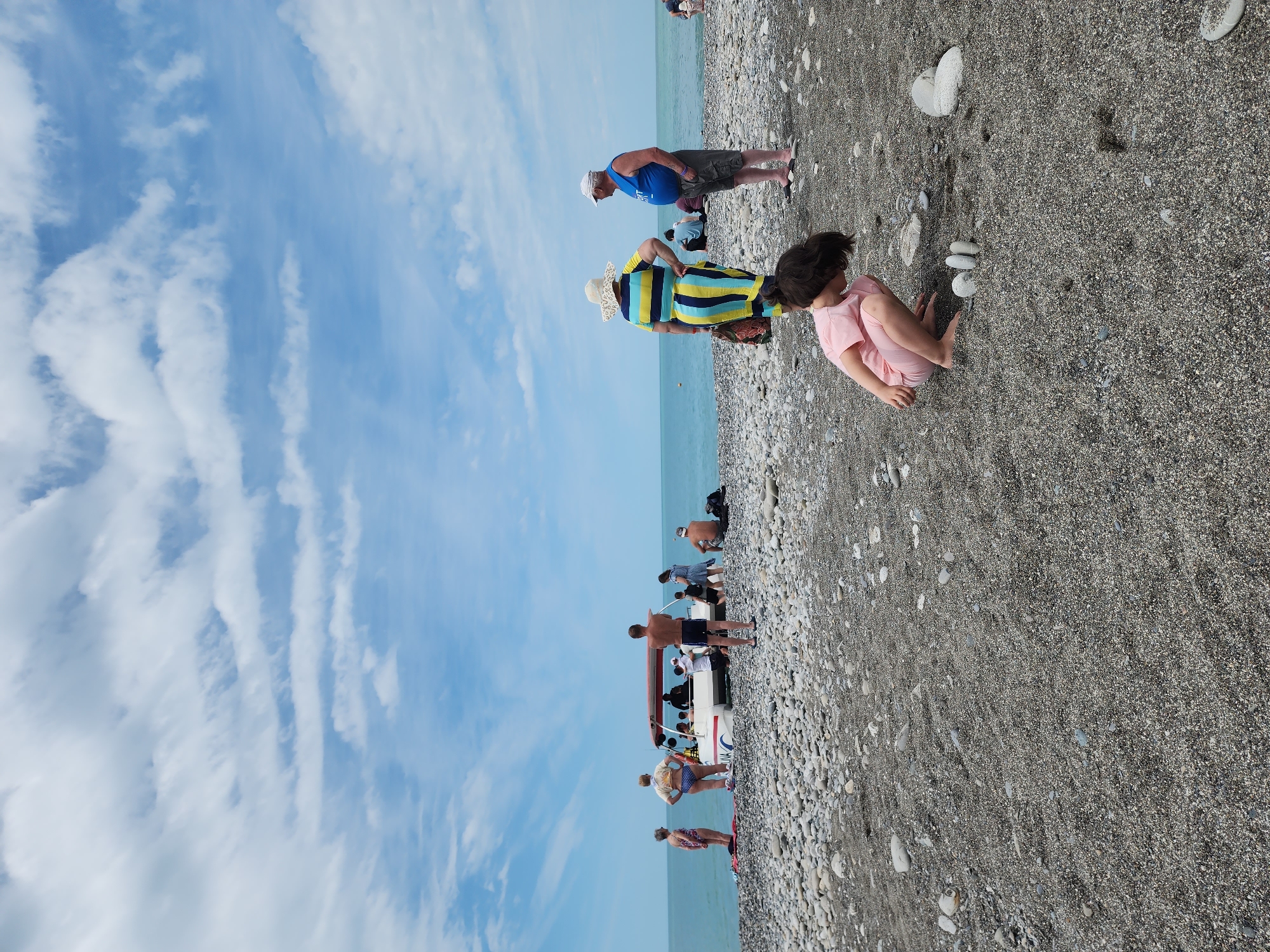 This photo shows a beach scene in Sochi, Russia. In the foreground, a child in pink clothing sits on a pebbled beach, playing with small rocks. Behind the child, an adult wearing a hat and striped clothing stands near another adult in blue. Further down the beach, there is a group of people standing near a small boat. The sea stretches into the distance, meeting a partially cloudy sky.