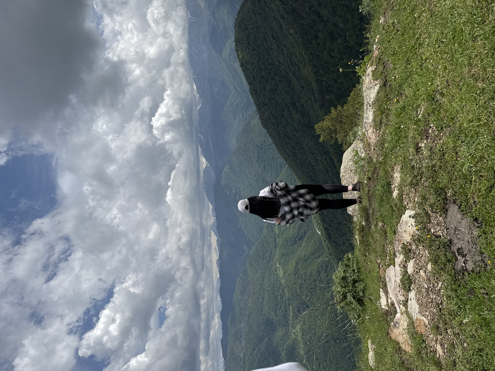 The image depicts a person standing on a rocky, grassy outcrop overlooking a vast, verdant mountain range. The person, viewed from behind, is wearing a white t-shirt, black leggings, a white baseball cap, and a plaid shirt draped over their shoulders. They are facing away from the camera, looking out at the expansive vista of densely forested mountains that recede into the distance, culminating in snow-capped peaks under a dramatic, cloudy sky. The location is identified as Nizhnyaya Teberda, Russia. The foreground consists of uneven terrain with rocks, sparse vegetation, and grass. The overall scene conveys a sense of awe and exploration amidst a majestic natural landscape.